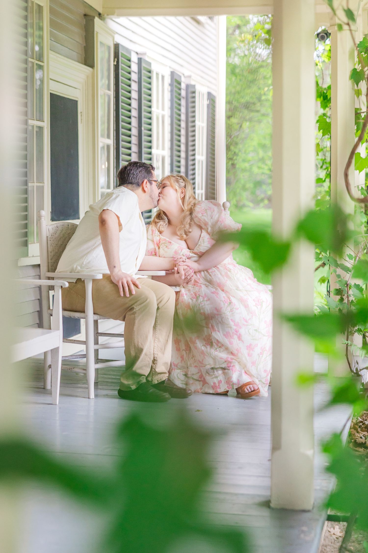 A couple embraces romantically on a white front porch surrounded by green foliage and white columns.