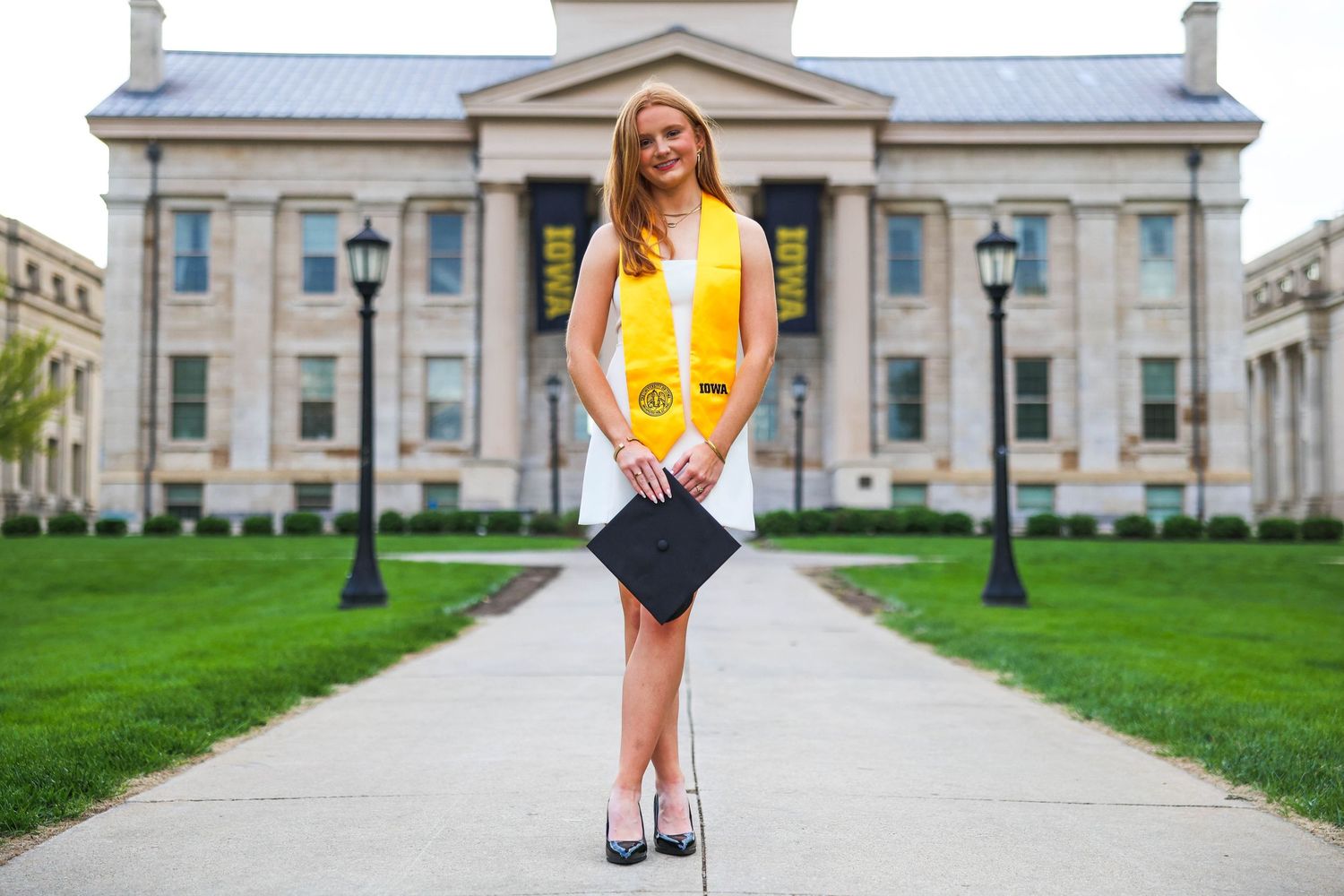 A graduate in yellow dress stands confidently on a grand university building's steps for a graduation photoshoot.