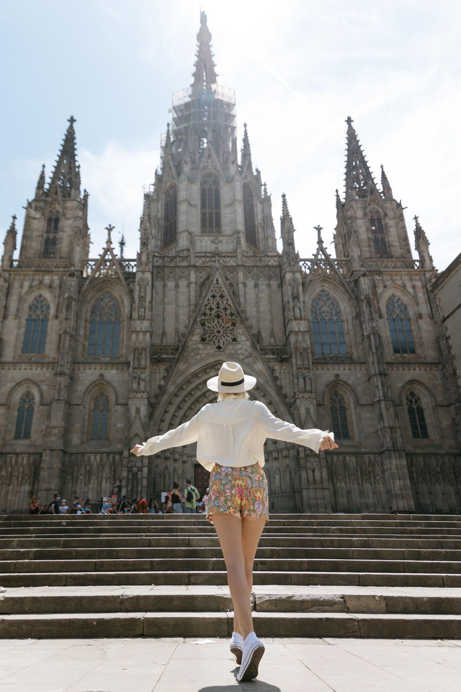 A person in a white dress and hat poses playfully in front of Barcelona Cathedral's Gothic architecture.