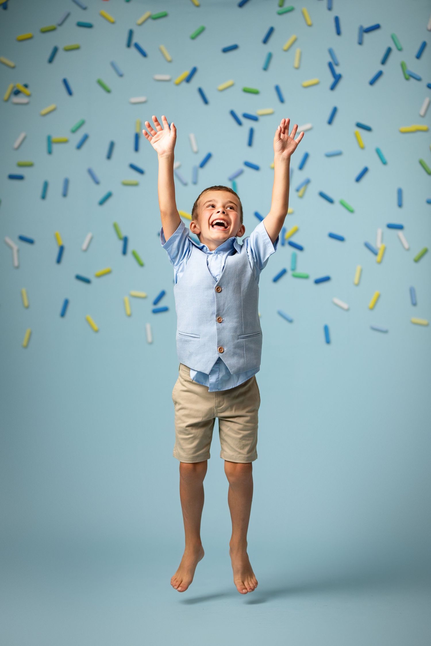 A small child holds a large gold number 3 balloon while wearing brown pants and a plaid shirt against a white background.