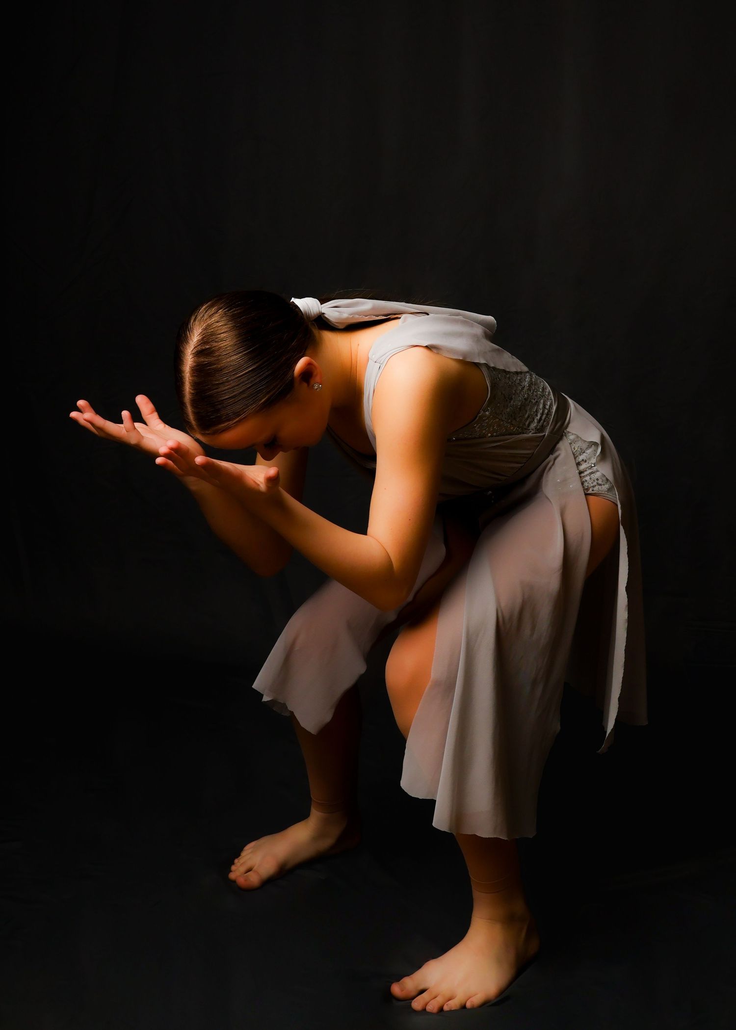 A dancer in a flowing white dress bends forward in an expressive pose against a black backdrop.