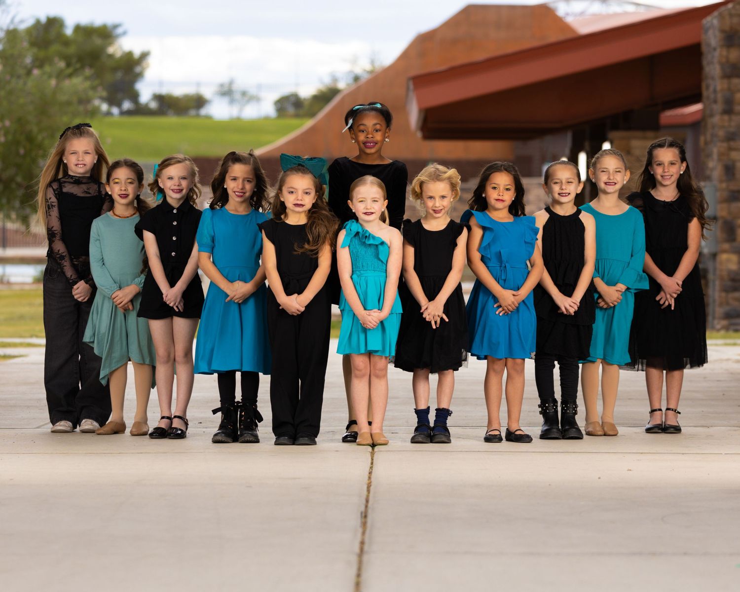 Young dancers lined up wearing turquoise and black dance outfits pose together outside a building on a sunny day.