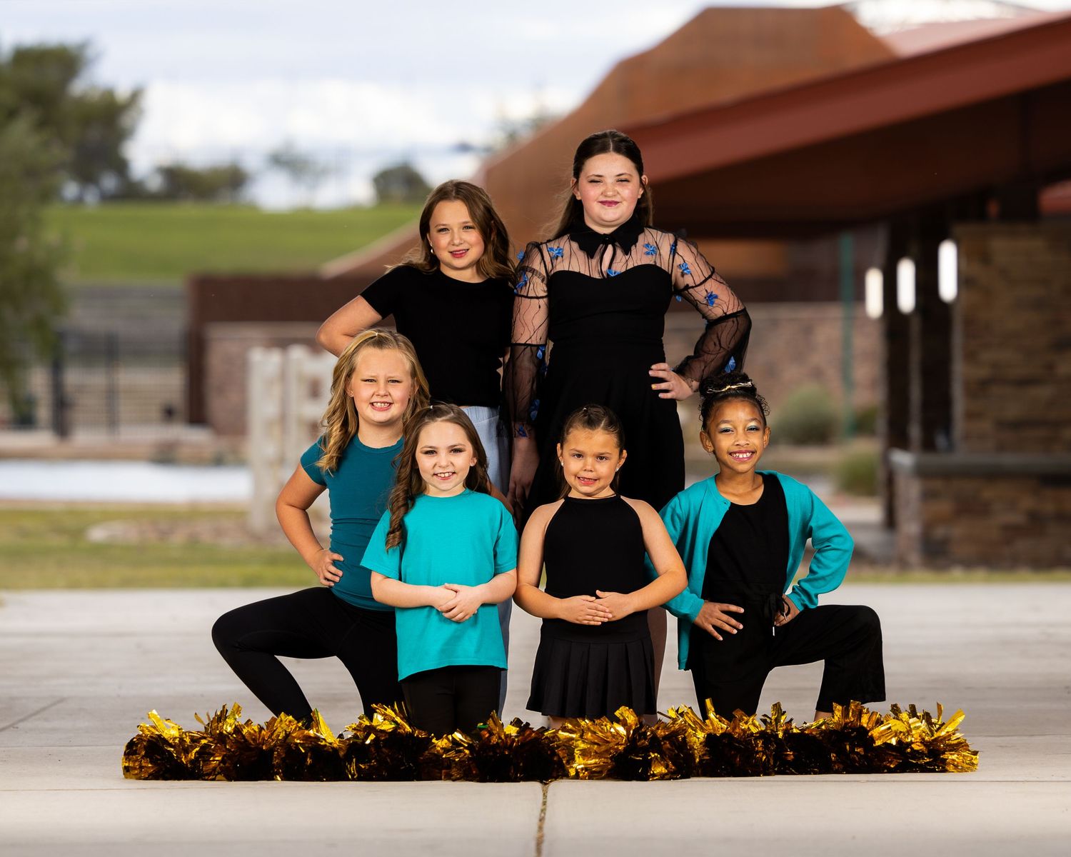 Dance group in matching turquoise and black outfits poses with gold pom poms on outdoor concrete surface.