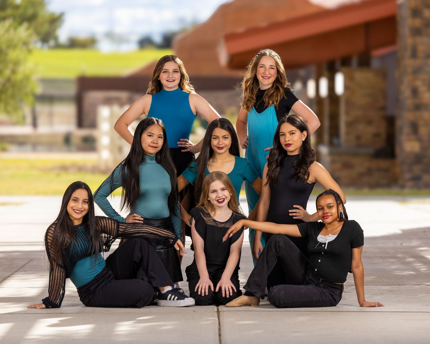 A dance team poses together in teal and black outfits on a sunny day outside a brick building.