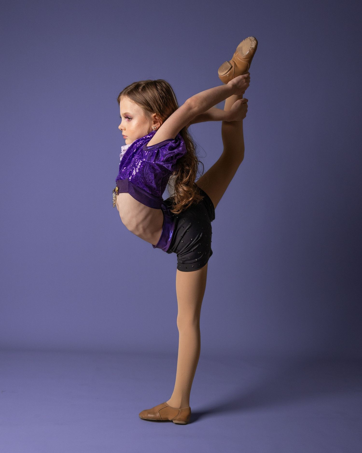 Young gymnast in purple leotard demonstrates impressive flexibility with high leg extension poses in studio setting.