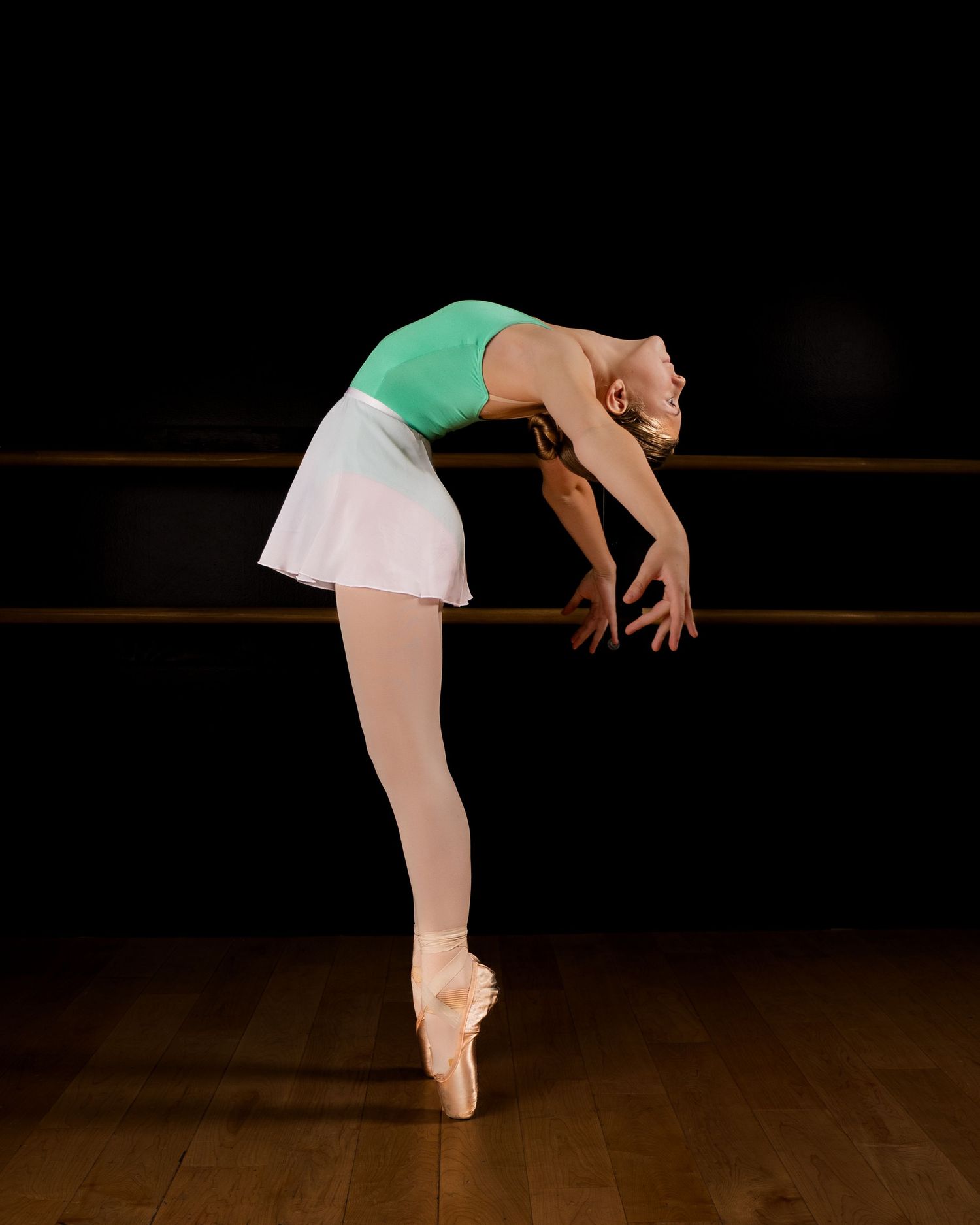 Ballet dancer in mint green leotard and white tutu performs graceful balance pose on dark stage.