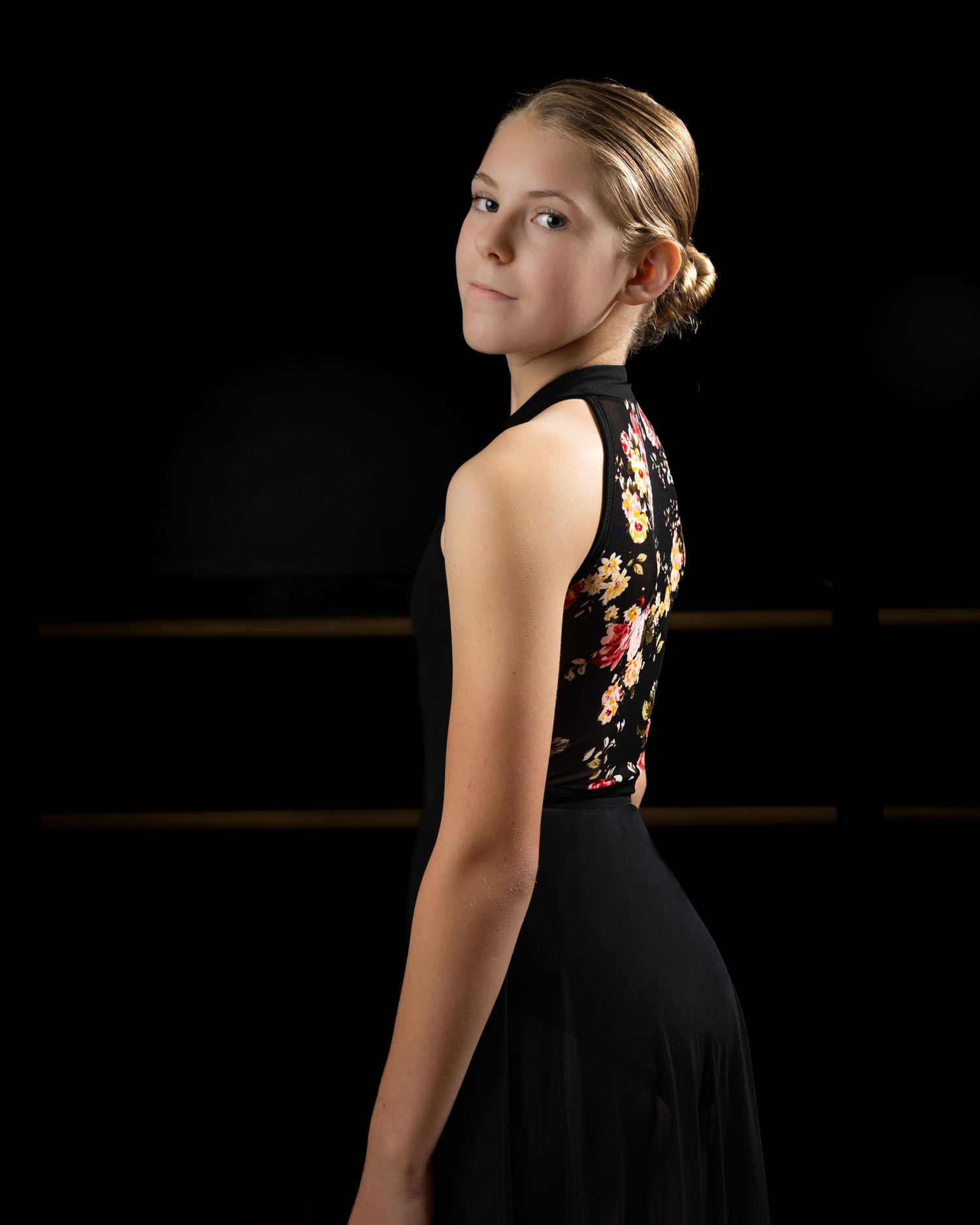 Woman in black dance costume with floral detail strikes elegant side pose against dark background.