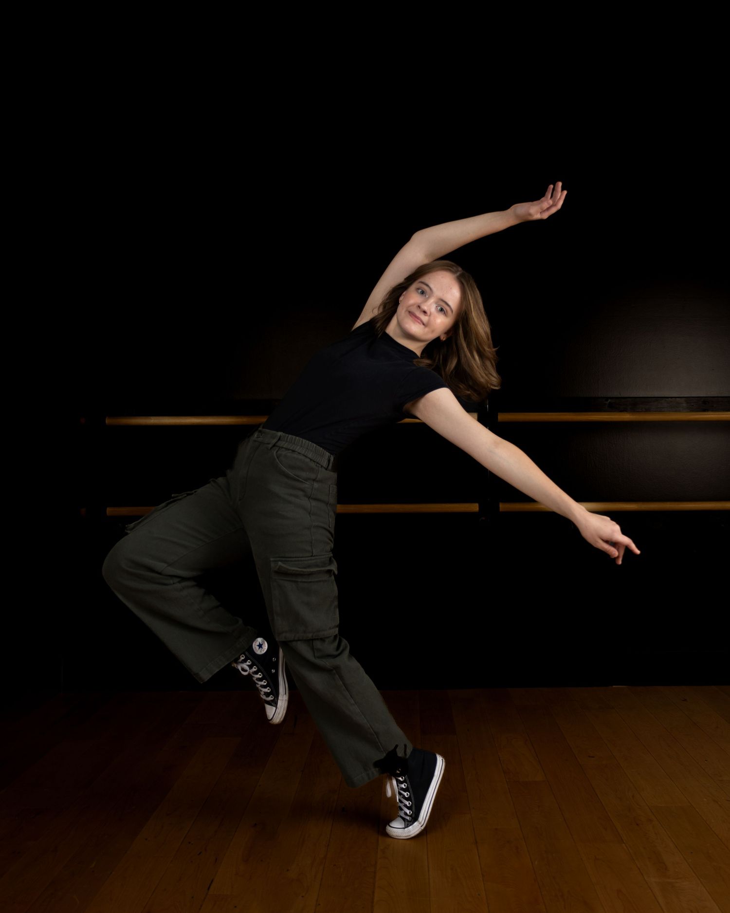 A dancer in dark pants performs a flowing movement sequence across a wooden dance floor in low lighting.