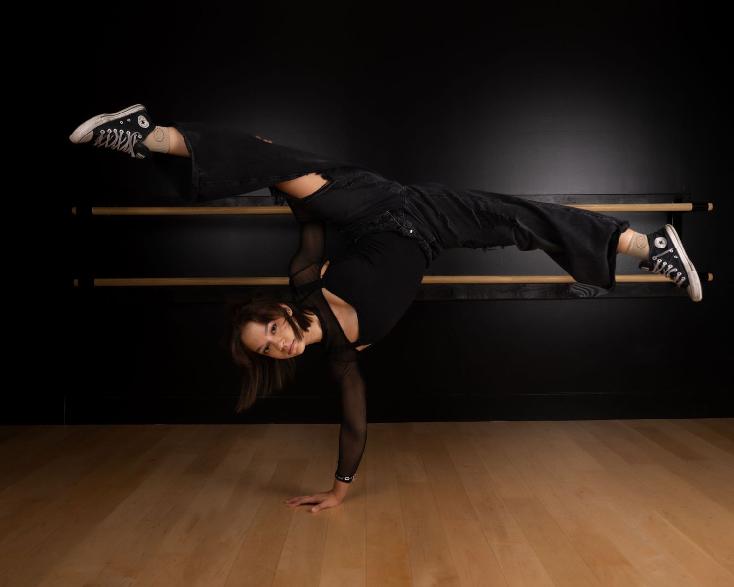 Dancer performing a dramatic horizontal balance pose on ballet barre against dark background in dance studio.