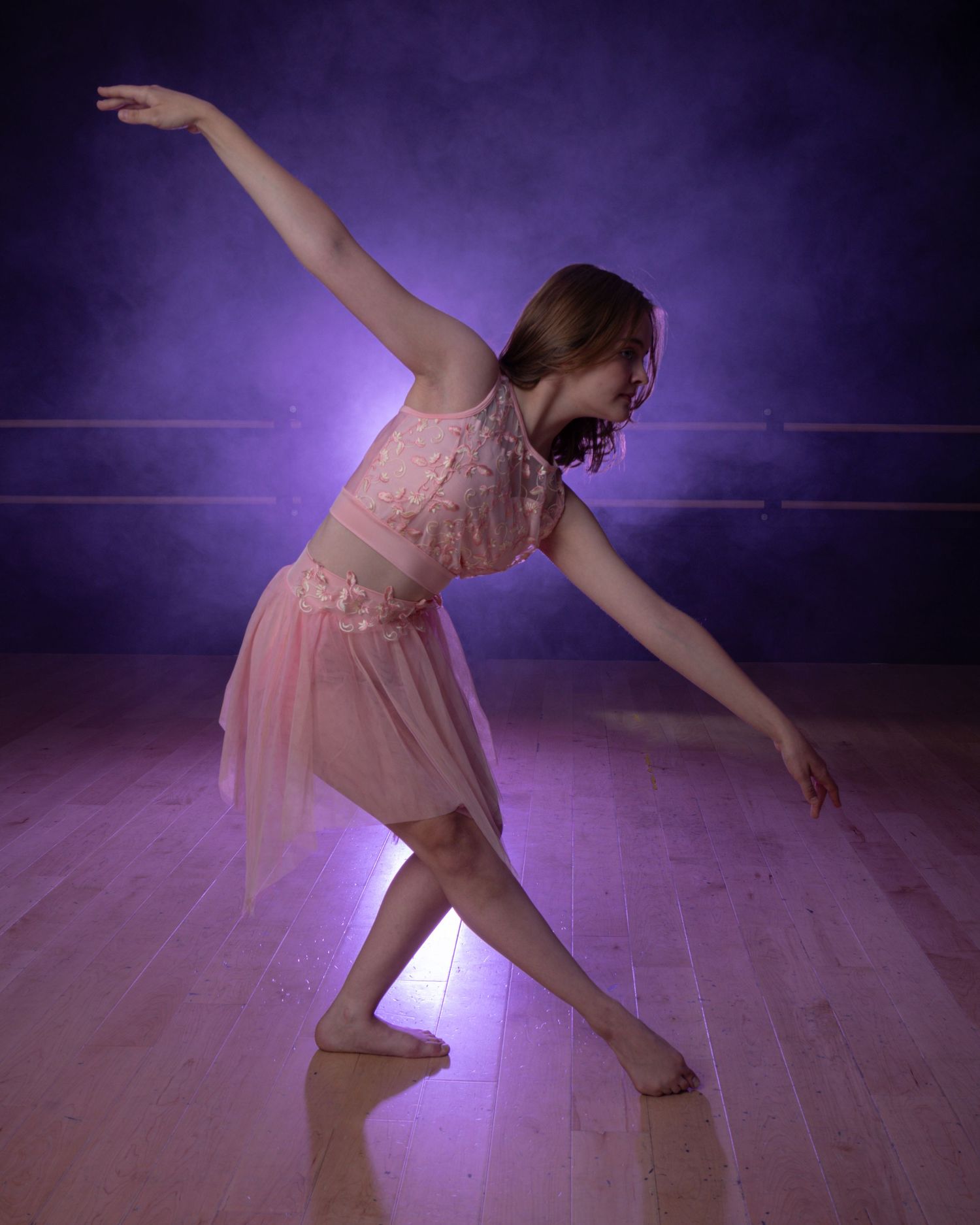Ballet dancer in pink dress practices graceful movements on dark stage under purple lighting.