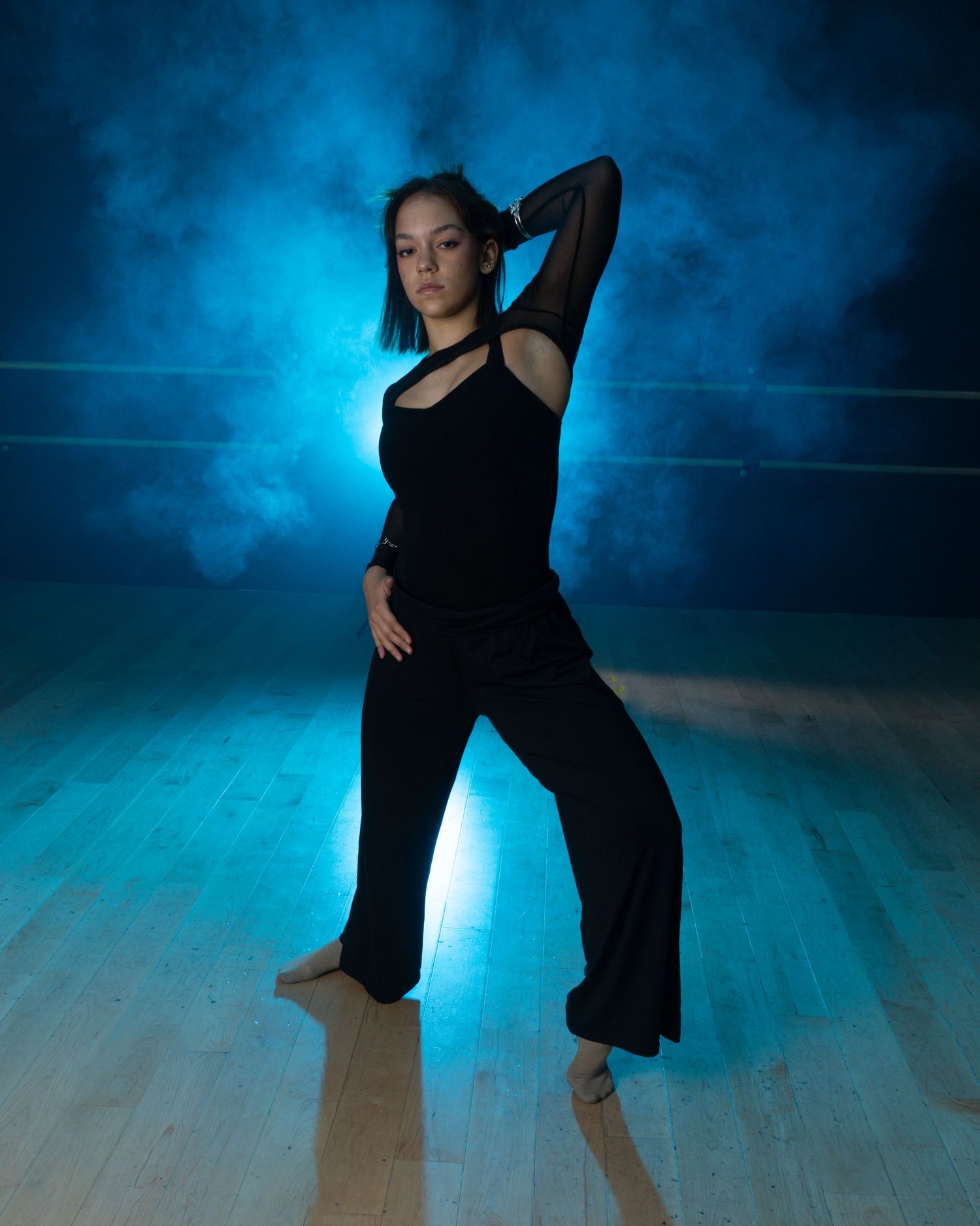 Dancer in black outfit strikes elegant pose against dramatic blue background on wooden dance floor.