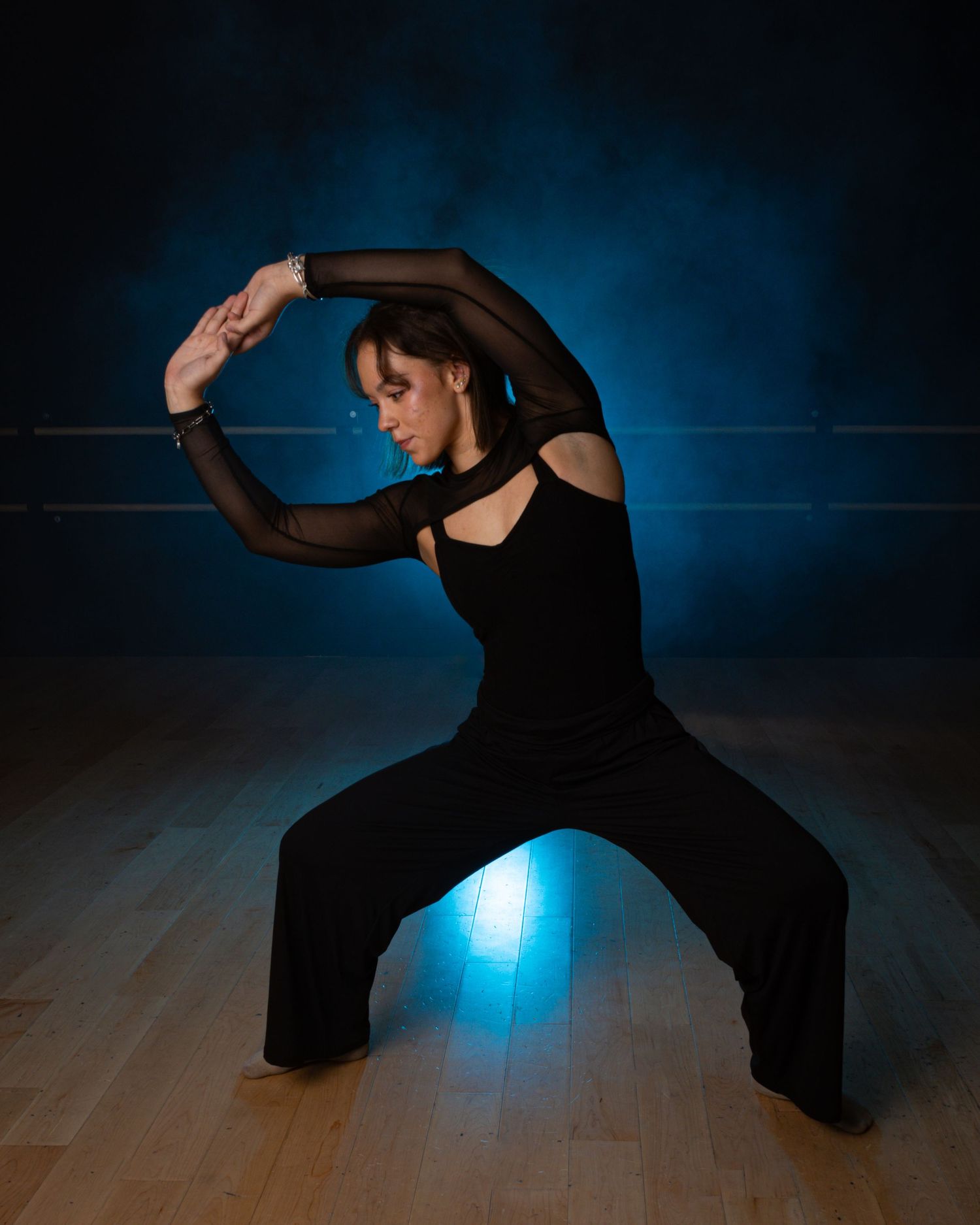 Dancer performs expressive movement in black attire against moody blue lighting on dance stage.