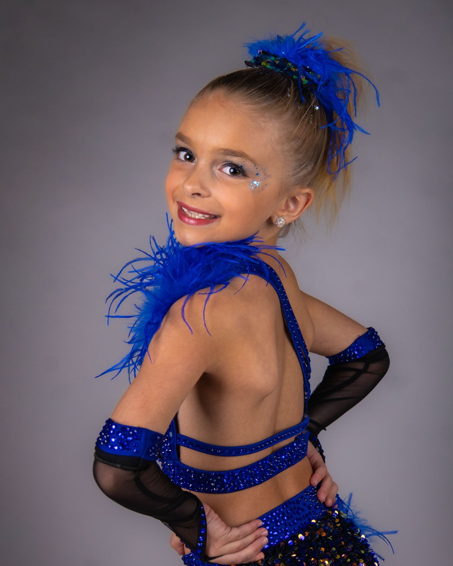 Dancer in sparkly blue and black performance costume with feather accents poses against gray background.