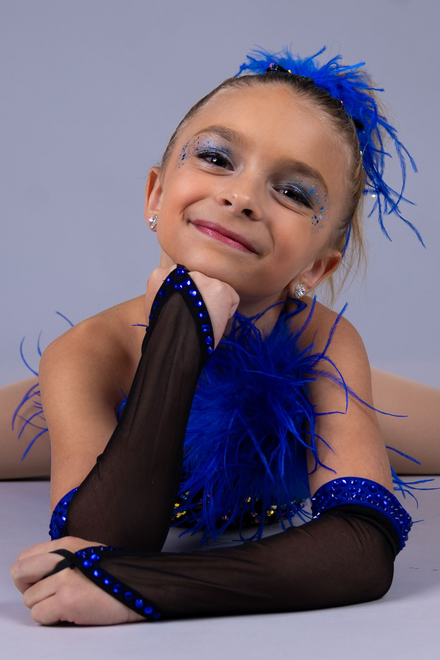 Dancer in blue feathered costume and black gloves performs expressive movements against light studio backdrop.