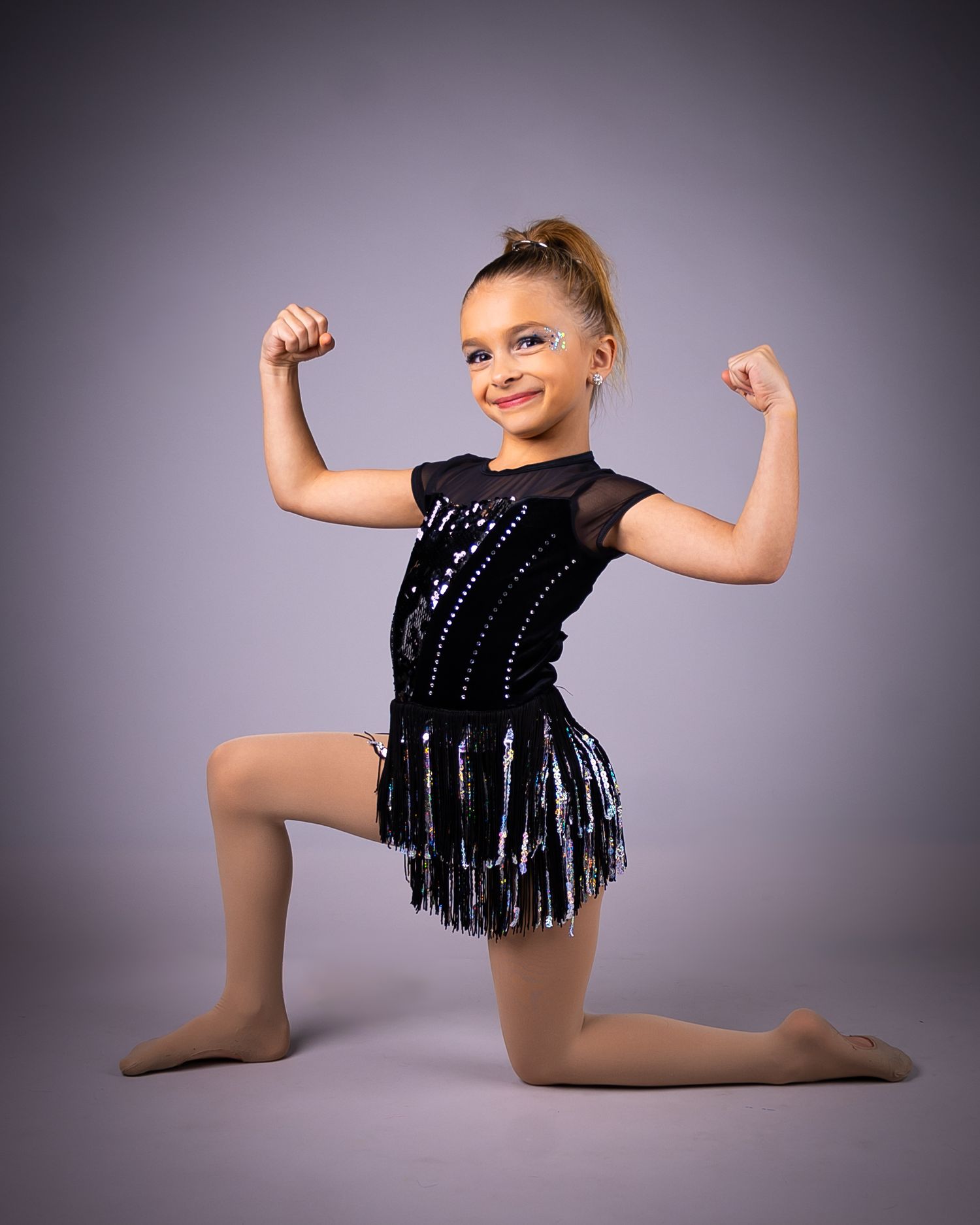 Young dancer in black sequined costume with fringe performs energetic pose on studio floor with grey backdrop.