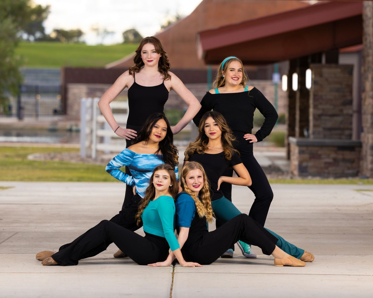 Dance group in black outfits with turquoise accents poses together on concrete in an artistic formation.