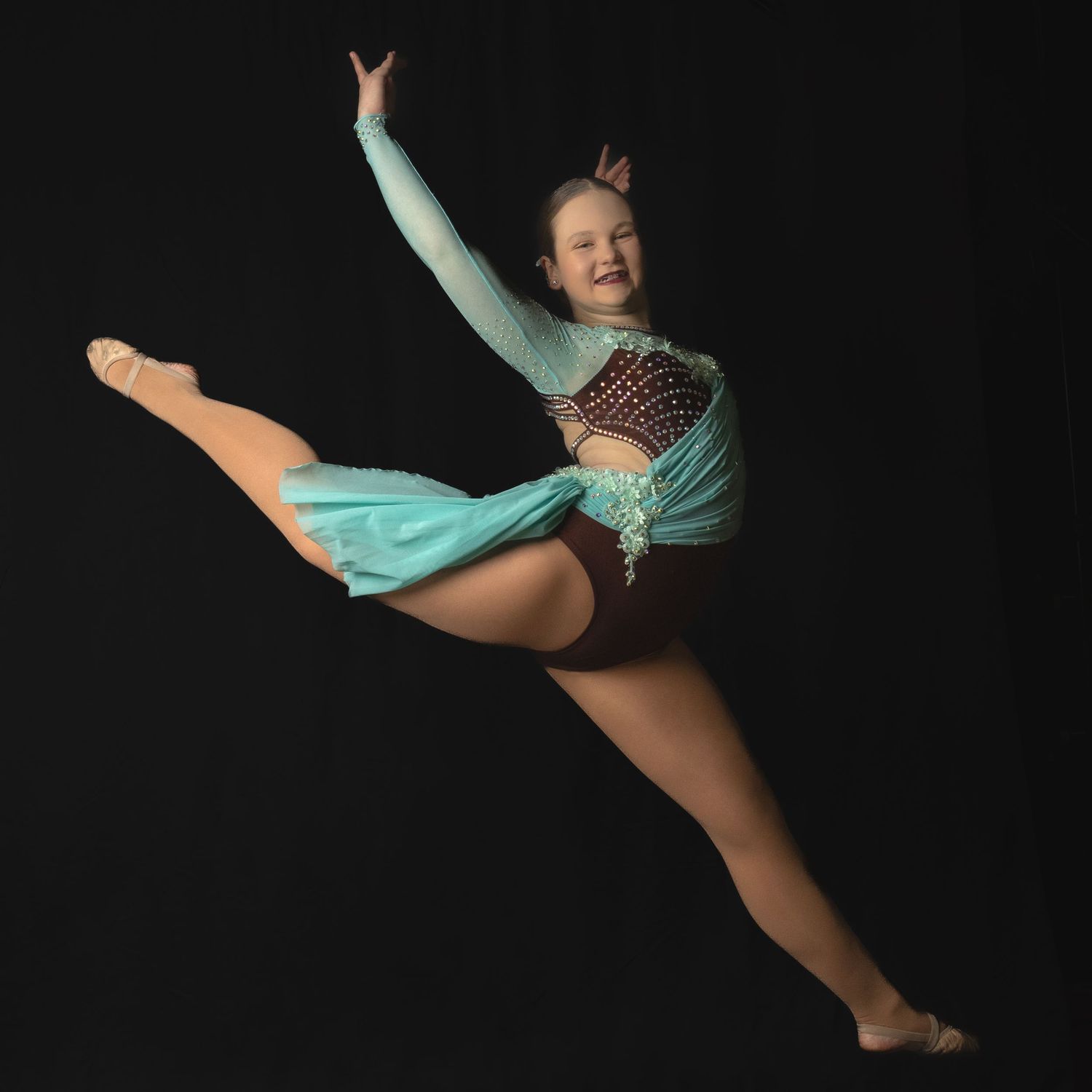 Young dancer in turquoise leotard performs graceful leap against black background in dramatic studio lighting.