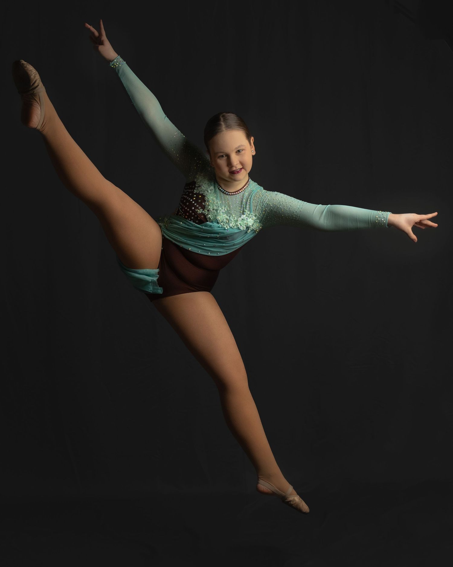 Graceful dancer performing dramatic split leap against dark background wearing teal leotard.