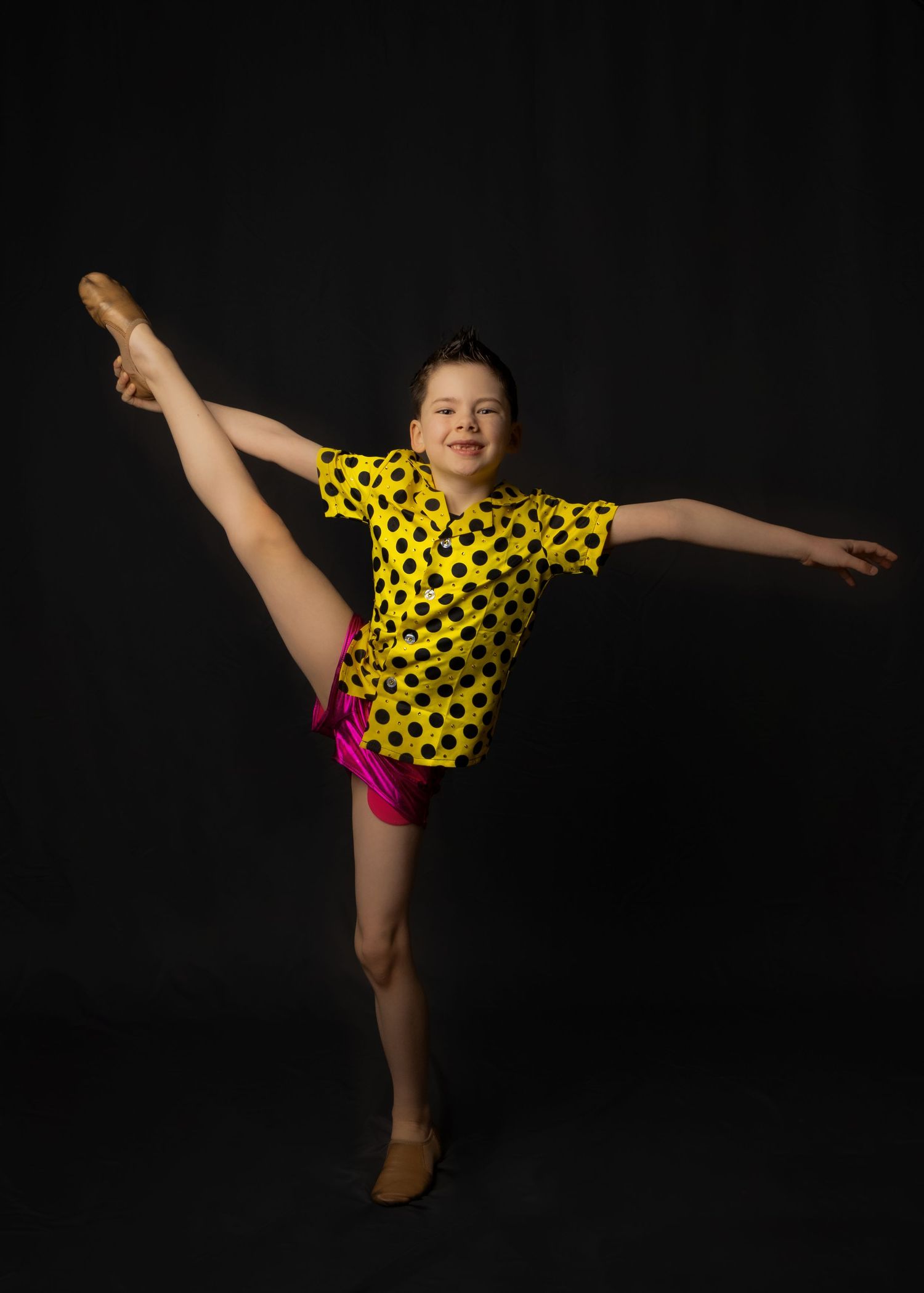 Young dancer in yellow polka dot top and pink shorts performs graceful high kick pose against dark background.