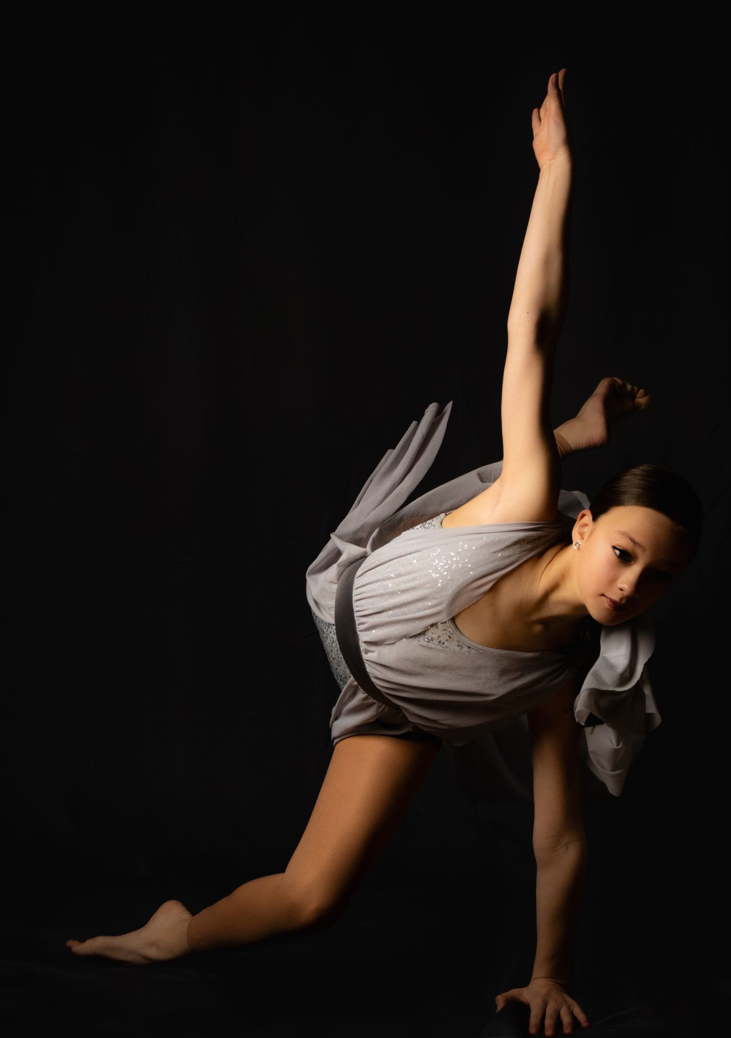 A dancer in a grey dress performs a dynamic pose against a dark background, stretching one leg upward gracefully.