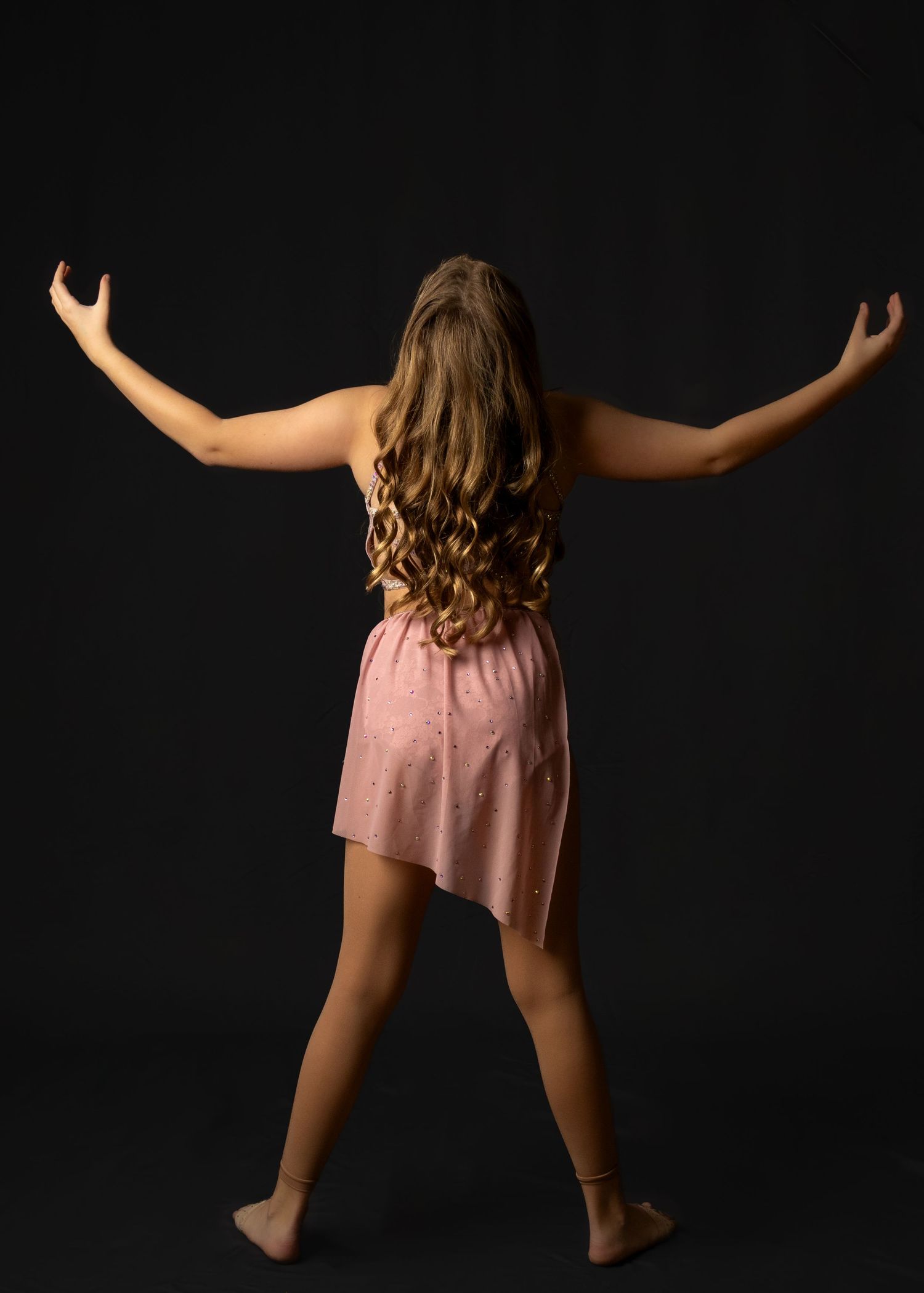 A person with long curly hair wearing a pink dress stands backwards with arms outstretched against a dark background.
