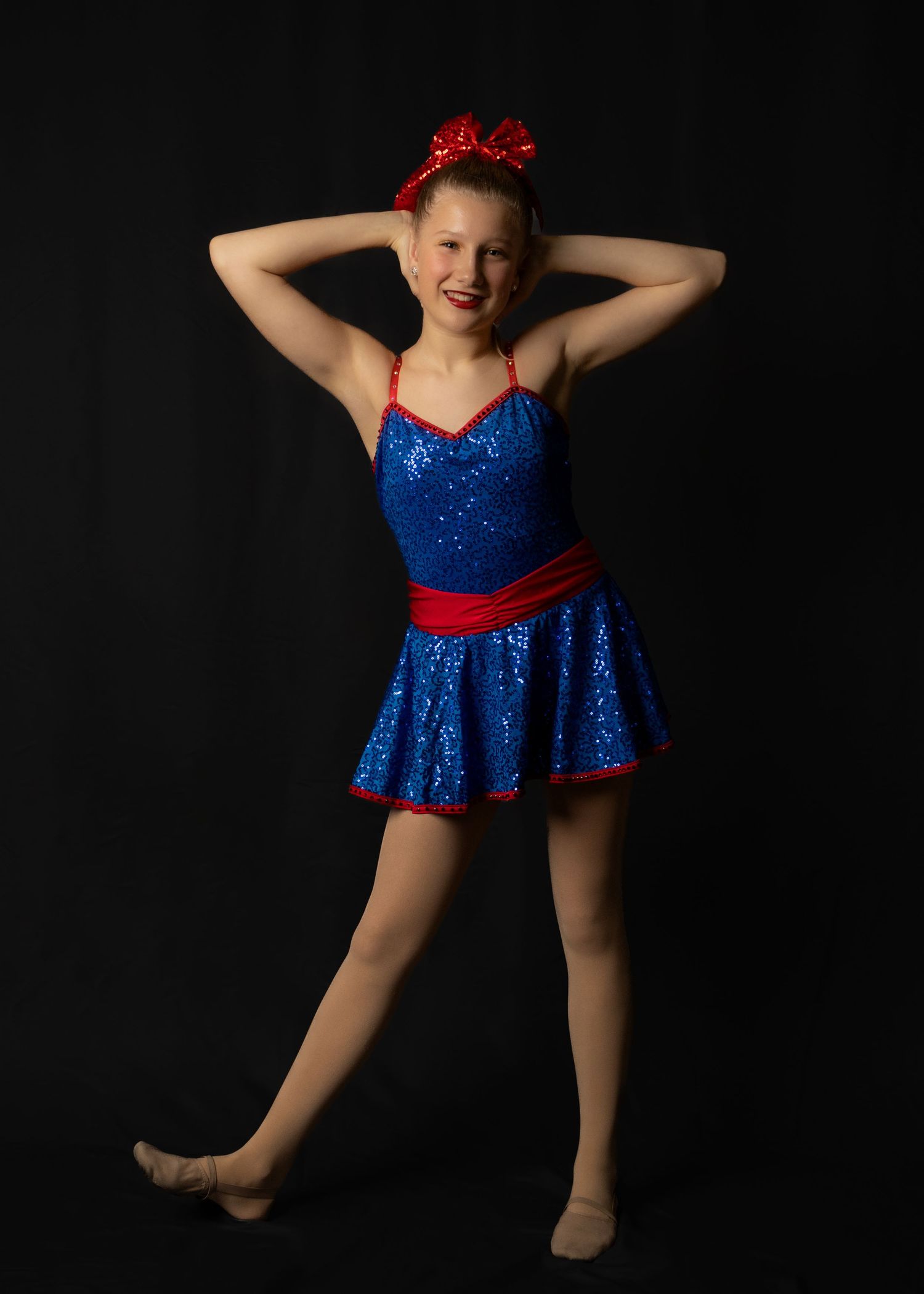 A dancer in a sparkly blue costume with red bow performs graceful poses against a black backdrop.