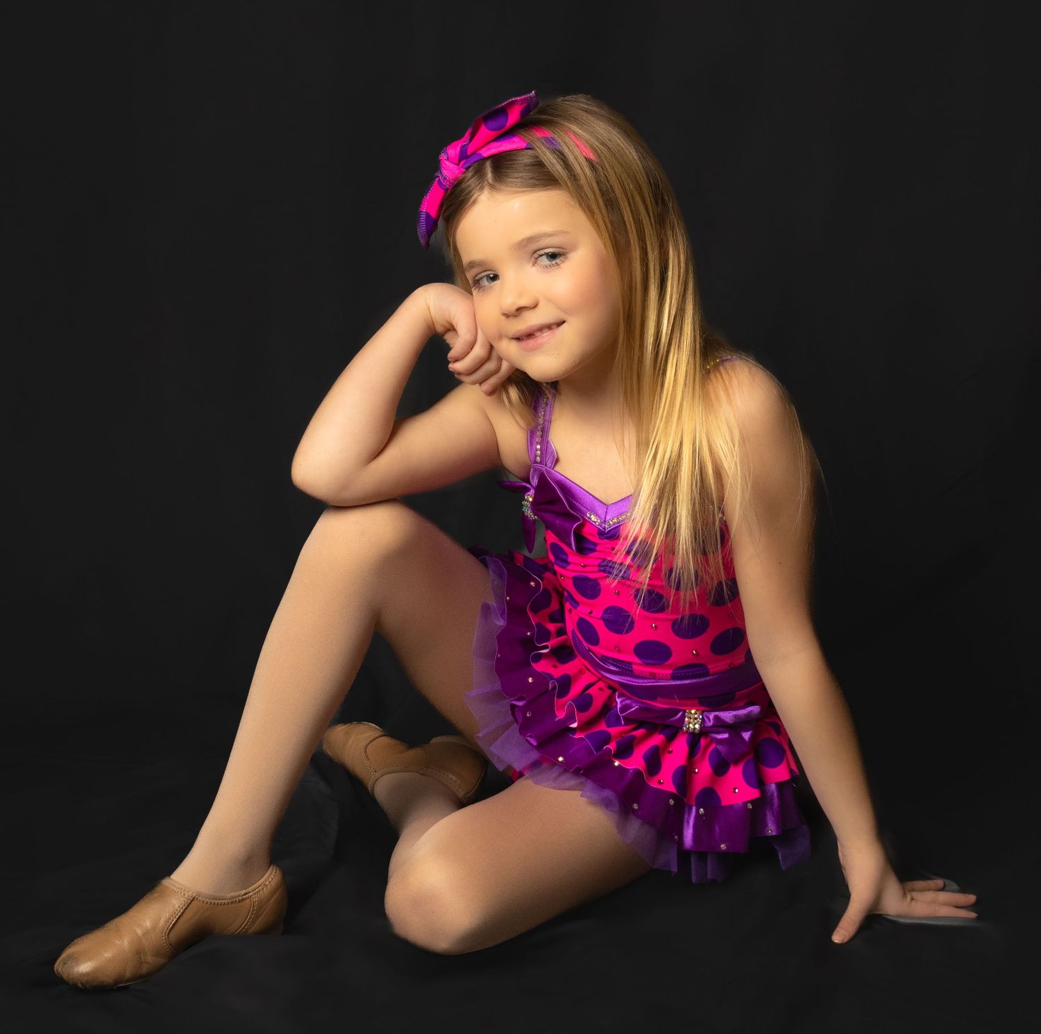 Young dancer in bright pink polka dot dance costume poses gracefully against dark background.