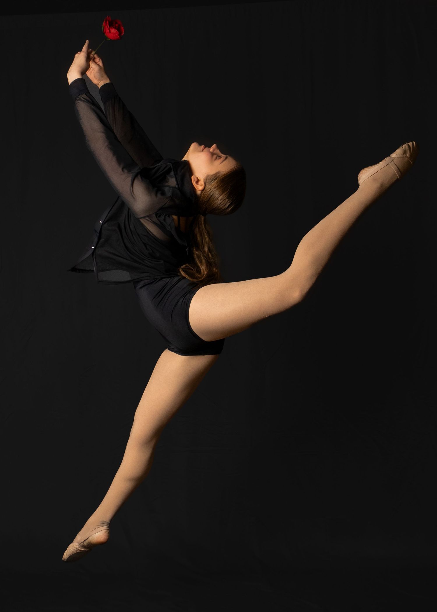 Dancer in black leotard performs dramatic split leap holding a red rose against dark background.