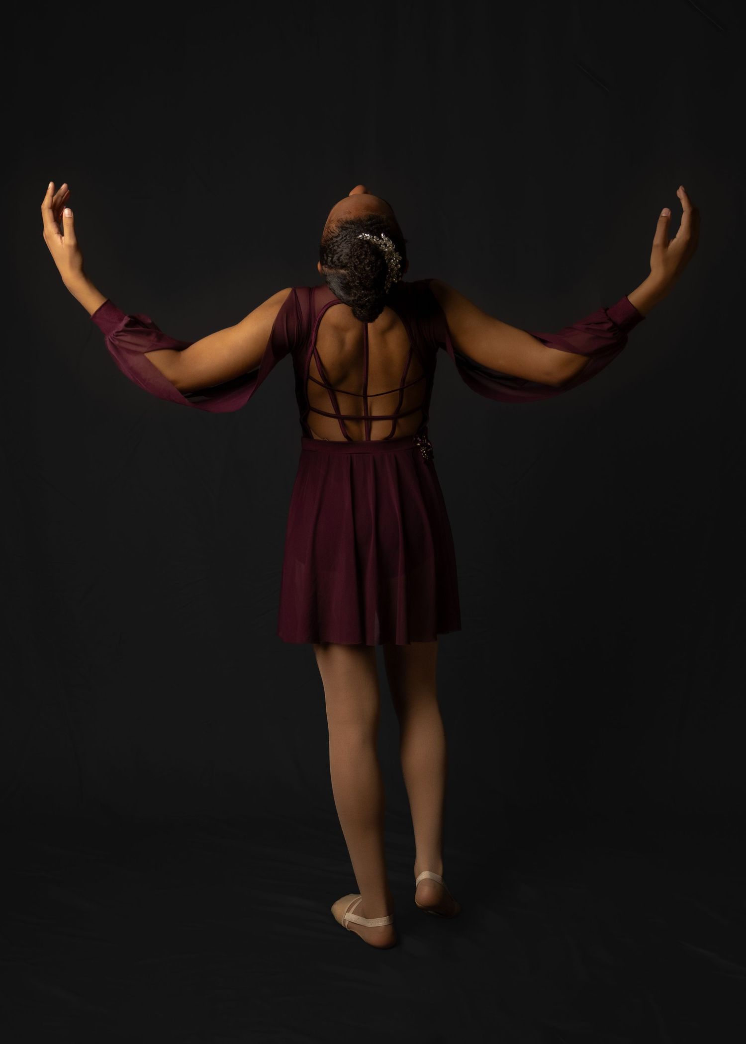 Dancer in maroon dress with crossed back design poses with raised arms against black backdrop.