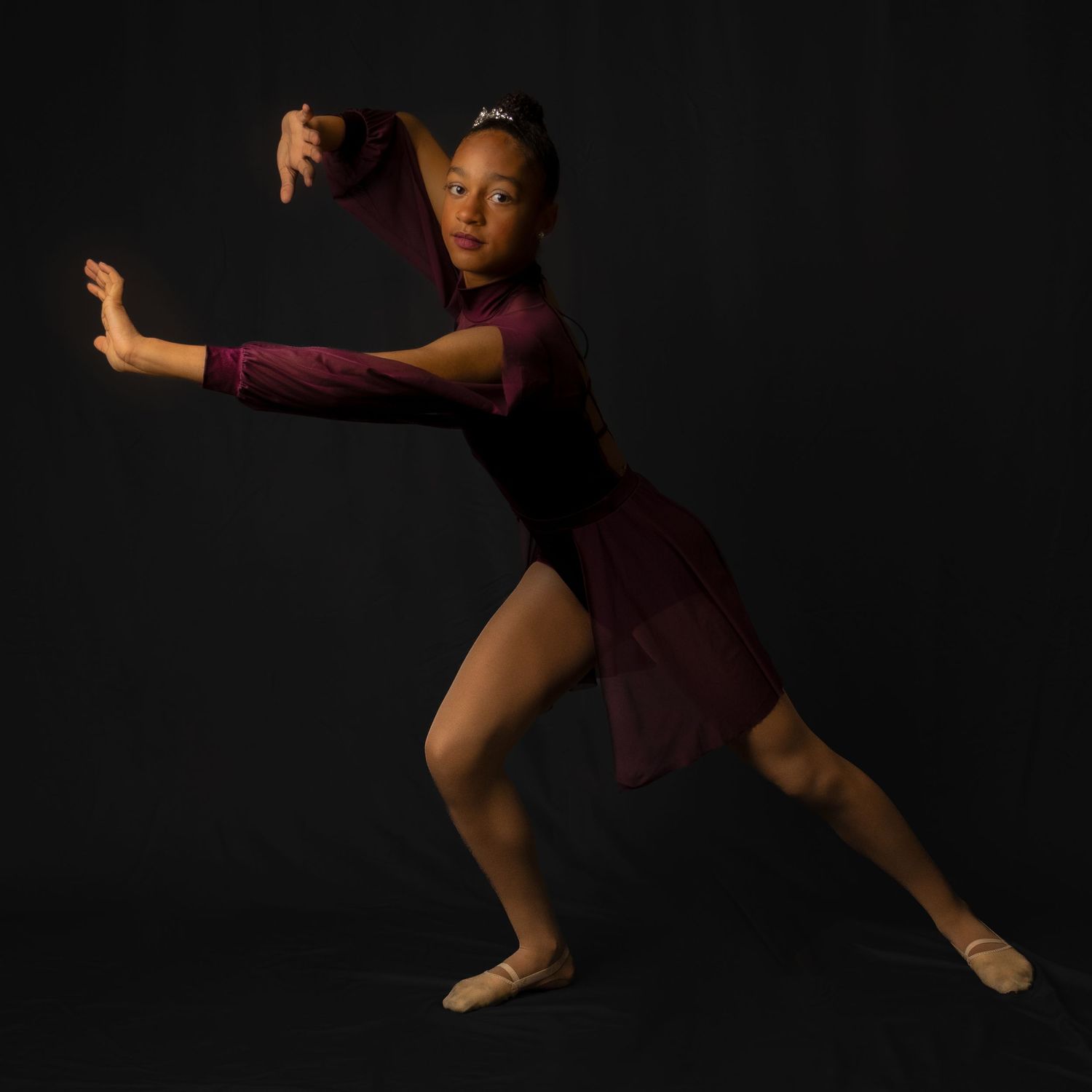 A dancer performs a graceful leap against a dark background while wearing burgundy dance attire.