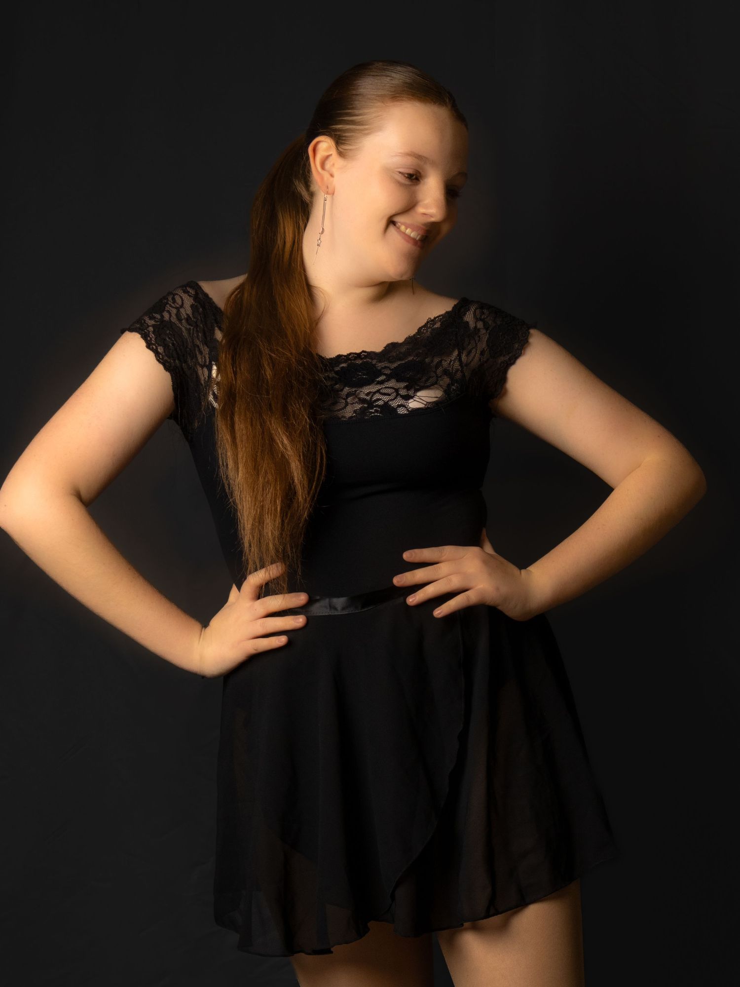A person in a black lace dress strikes a confident pose against a dark studio backdrop.