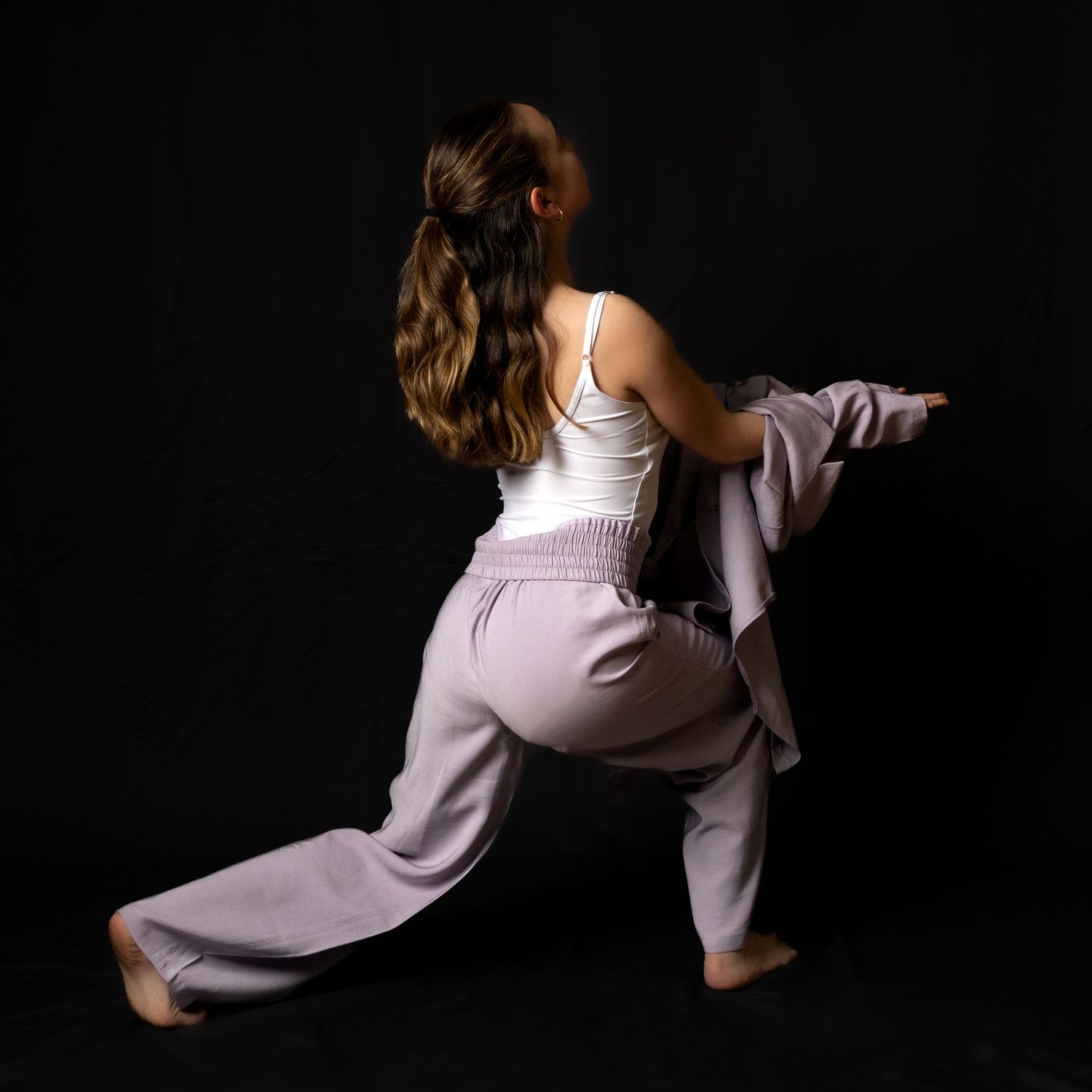 Figure in white tank top and loose gray pants strikes dynamic dance pose against dark background.
