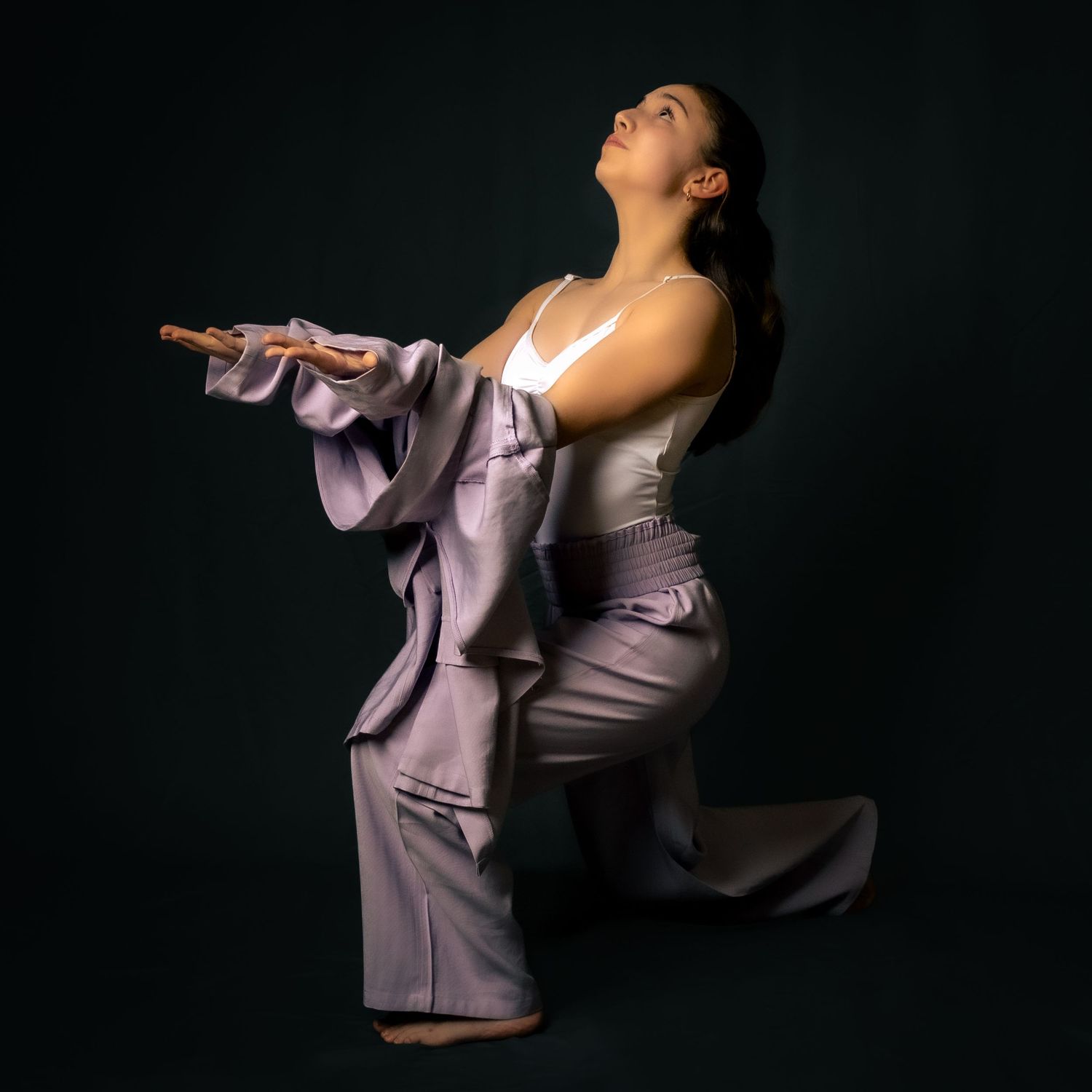 A dancer in a silk lavender outfit strikes a dramatic pose against a dark background, leaning back with flowing sleeves.