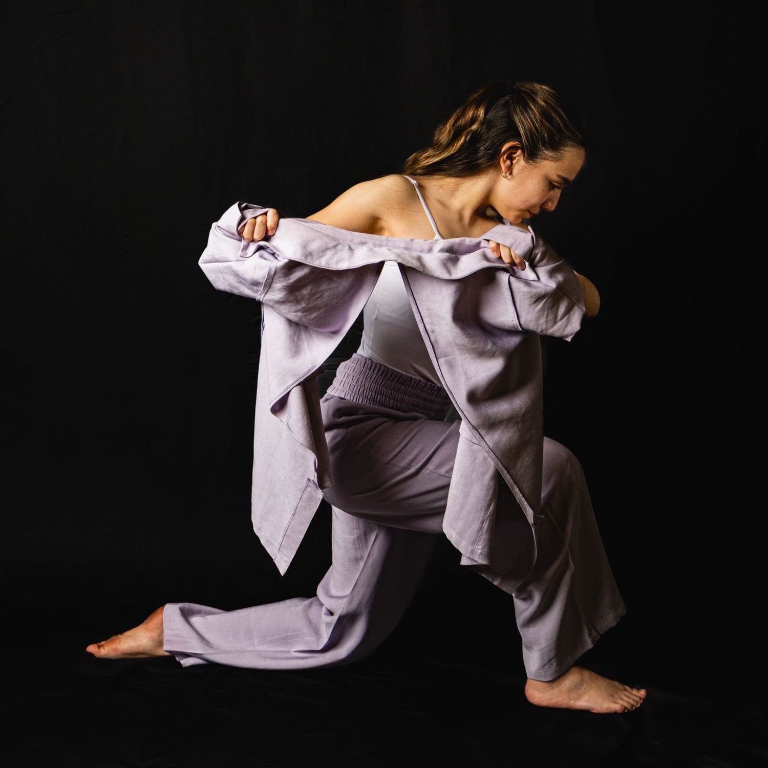 Contemporary dancer in lavender outfit performs dynamic movement against dark background with dramatic lighting.