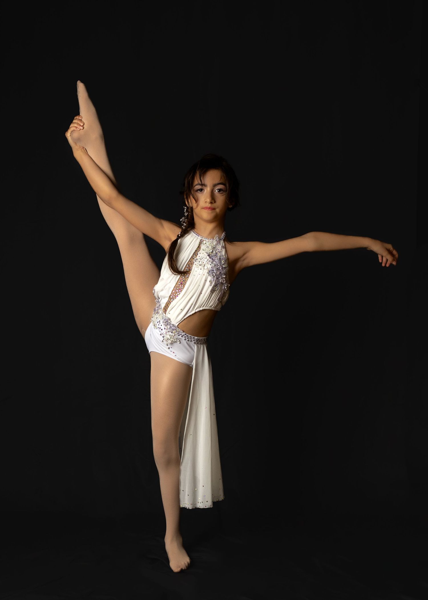 A dancer in a white sequined costume performs a high leg extension pose against a black background.