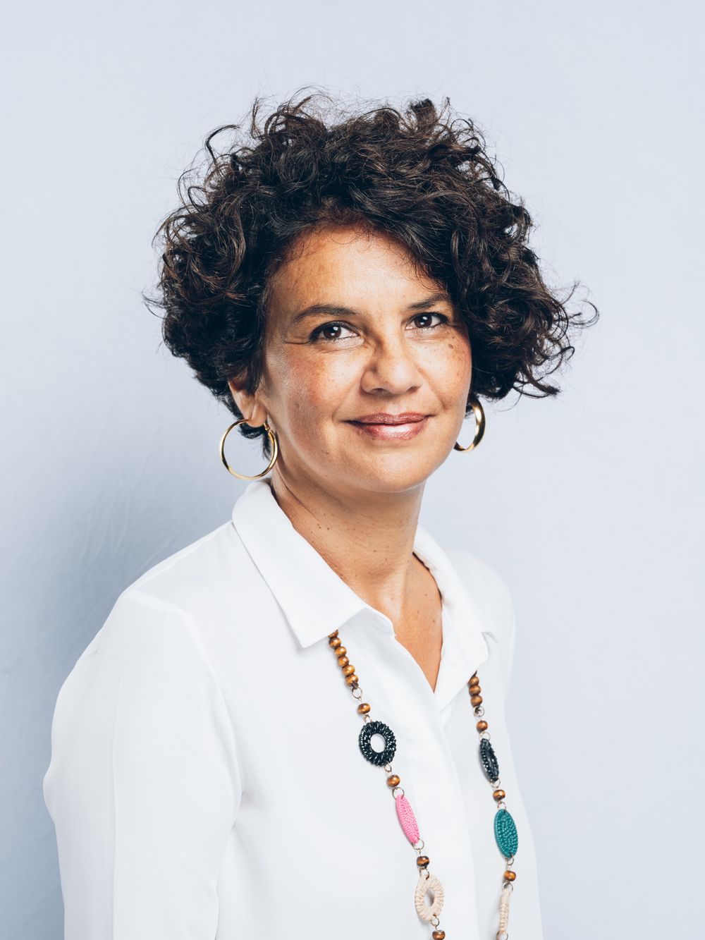 Woman with curly hair wearing white blouse and colorful beaded necklace with hoop earrings smiling confidently.