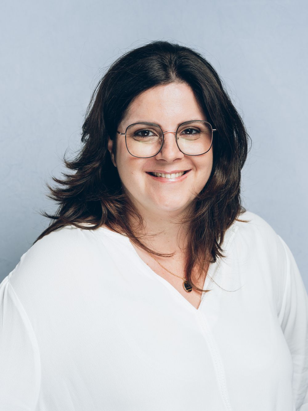Smiling woman with shoulder-length dark brown hair and round wire-rimmed glasses wearing a white blouse against a light blue background.