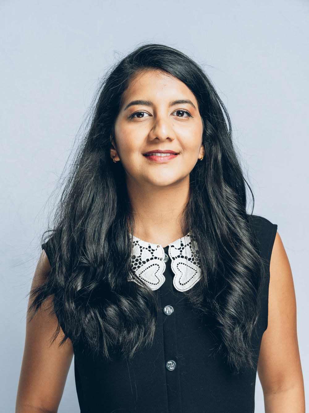 Young woman with long dark hair smiling, wearing a black sleeveless top with white lace collar detail.