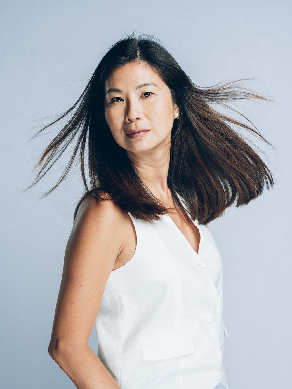Woman in white sleeveless top with flowing dark hair posed against light blue background.