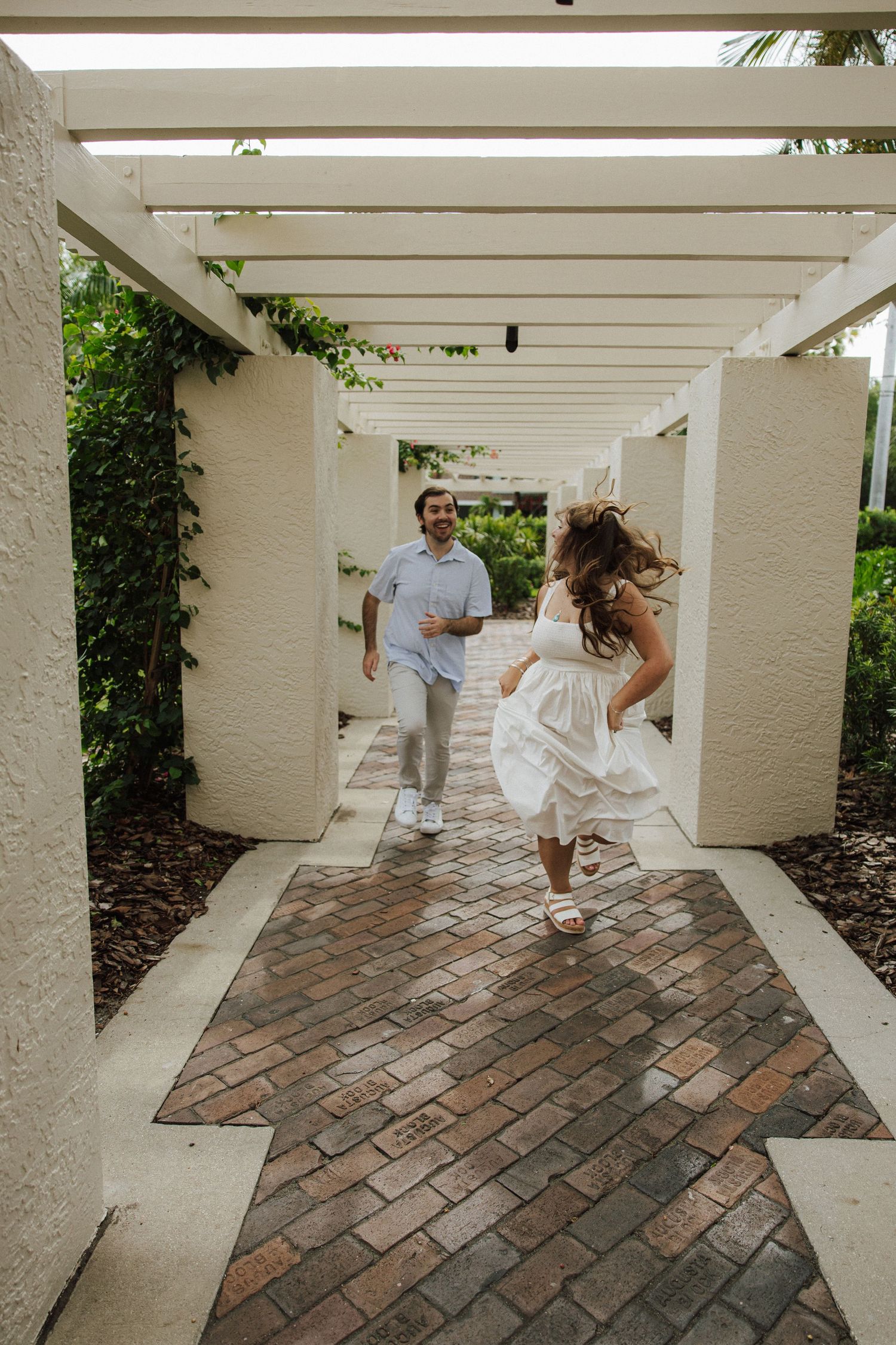 A couple running playfully down a covered brick walkway with white columns and vines growing along the corridor.