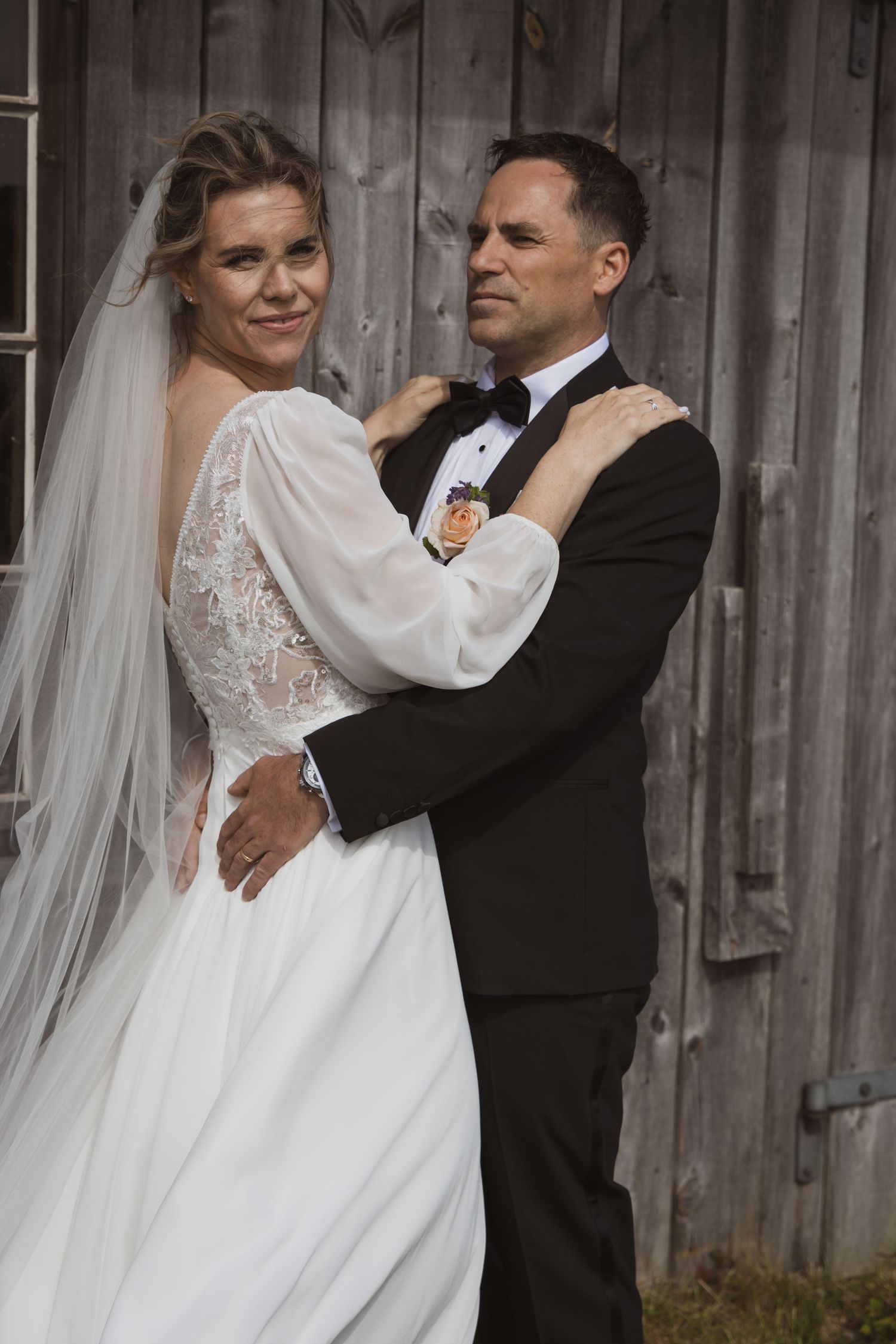 Bride and groom embrace tenderly in front of rustic wooden barn door in elegant wedding attire.
