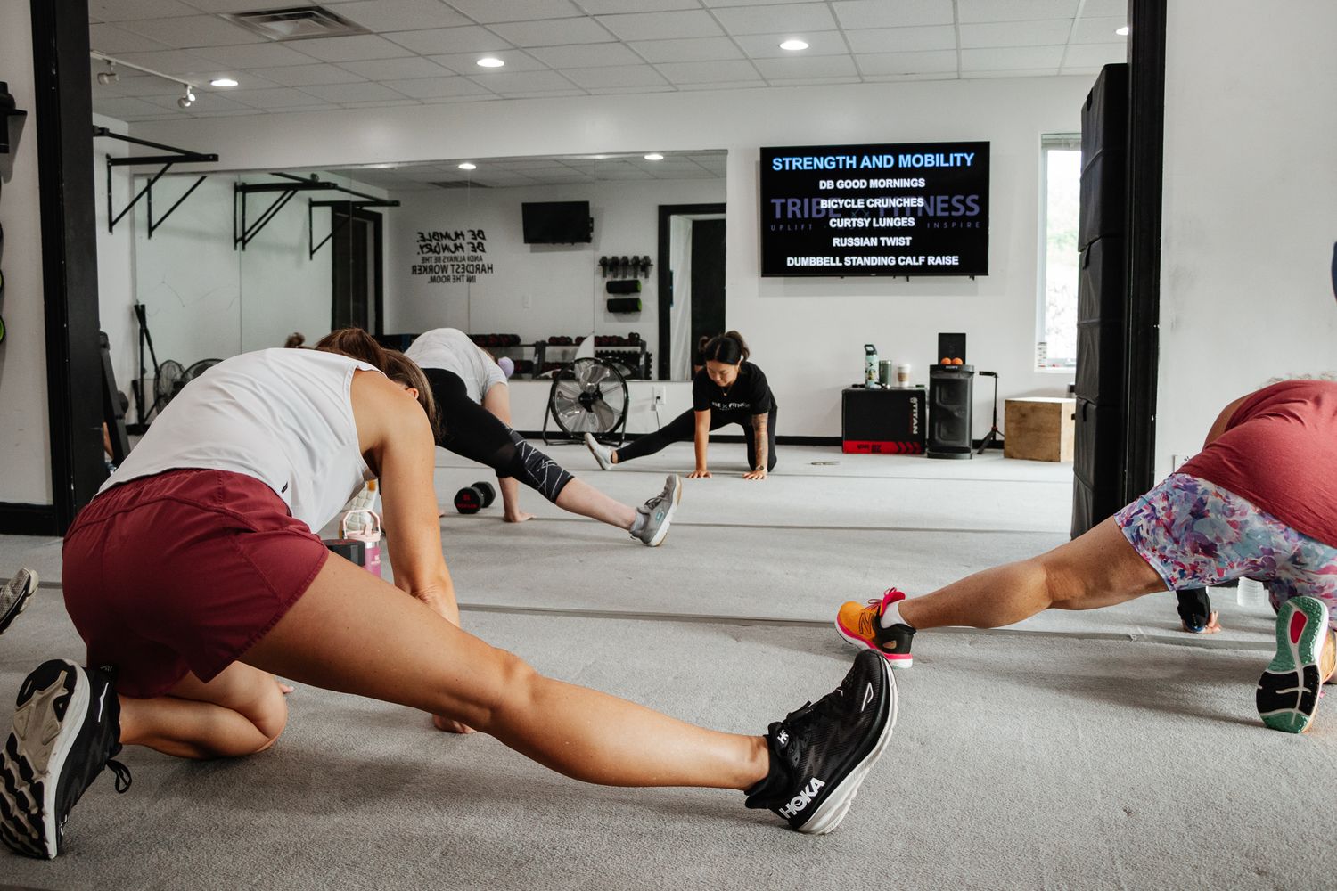 Group stretching session in a gym, participants lunging while instructor leads at the front near a mirror wall.