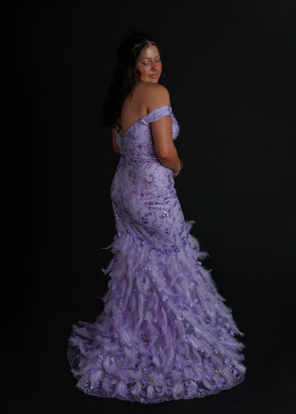 An elegant purple crushed velvet gown with off-shoulder design and dramatic feathered train against a dark background.
