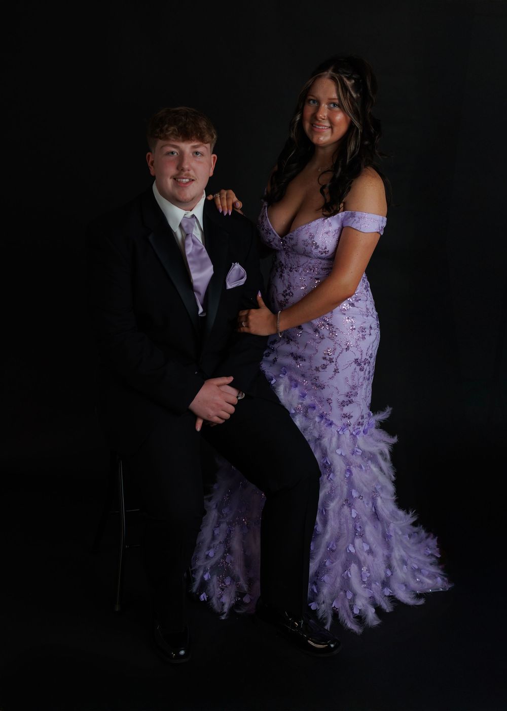A couple poses in formal attire at a prom or dance, with a lavender gown and black tuxedo against a dark backdrop.