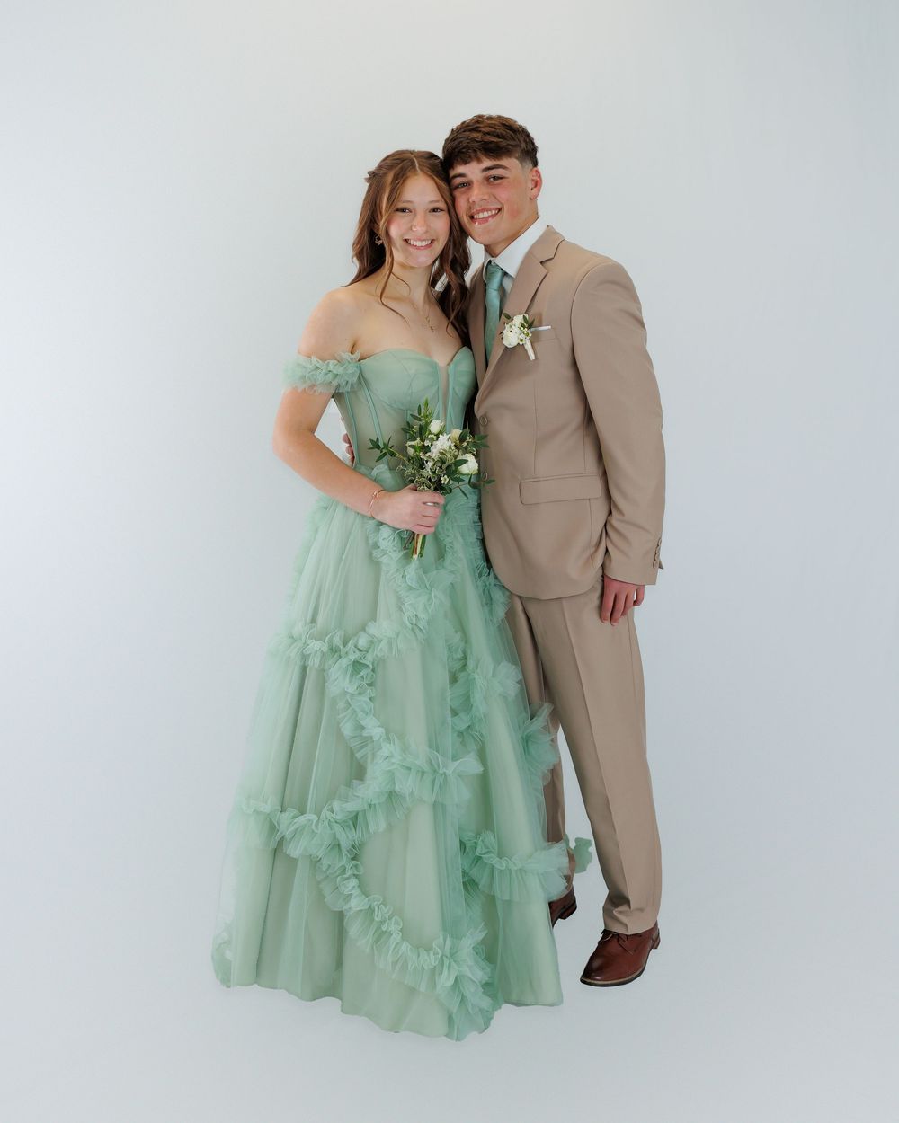 A couple poses together against a white background, wearing a mint green gown and tan suit for a formal occasion.
