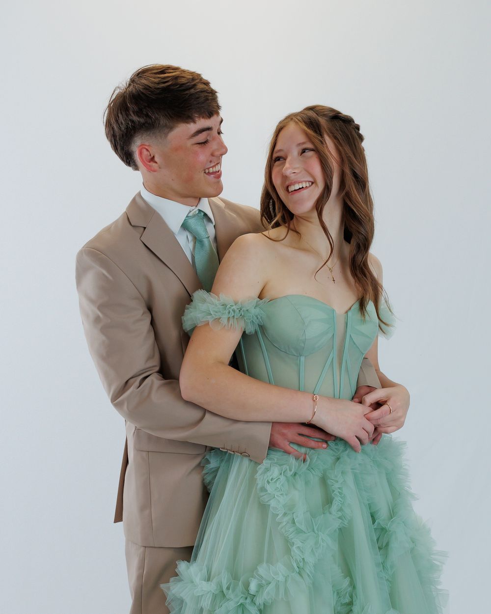 A couple in formal attire, with him in a tan suit and mint tie and her in a seafoam green tulle gown, share a loving moment.