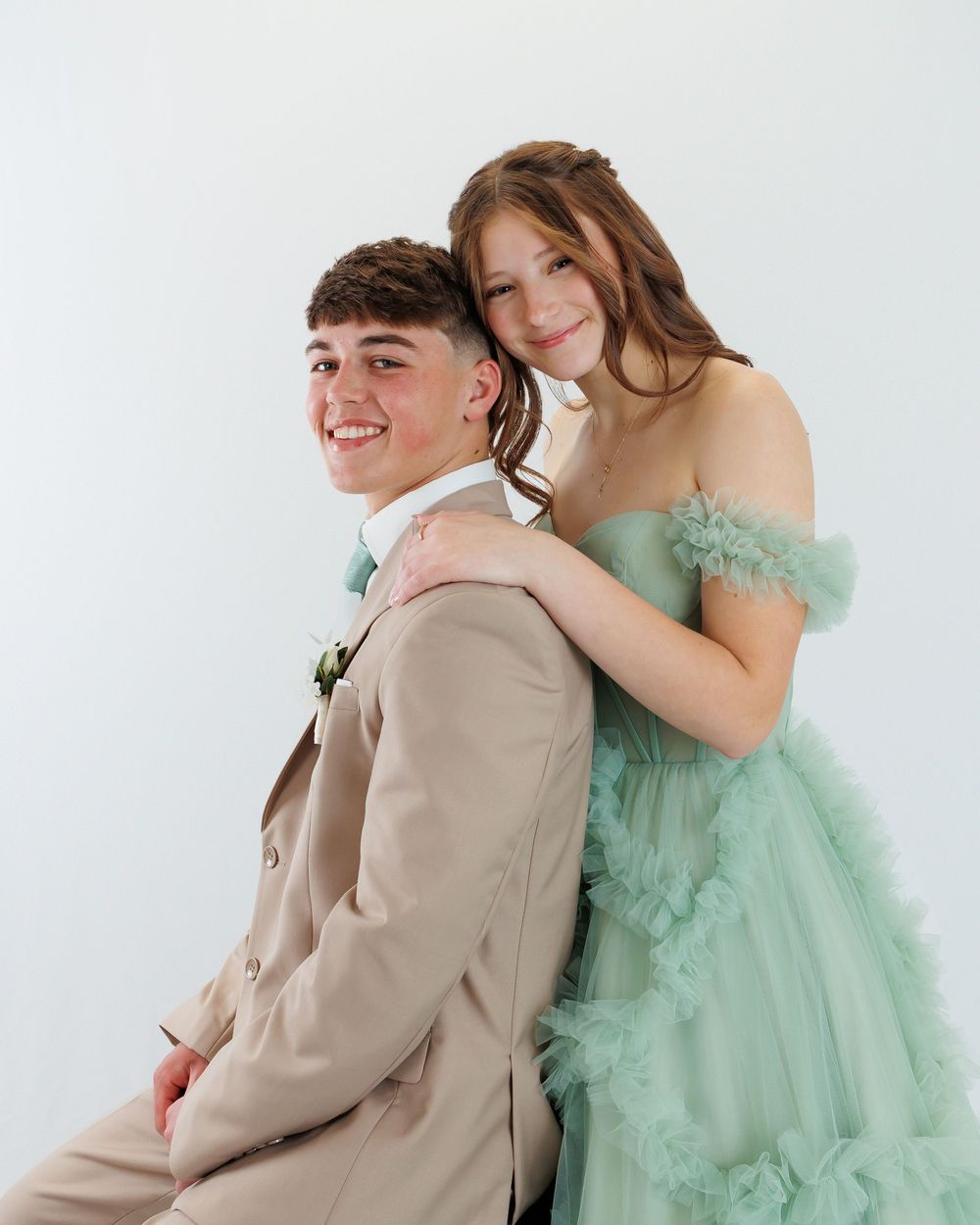 A couple poses together against a white backdrop, with one wearing a mint green tulle gown and the other in a beige suit.