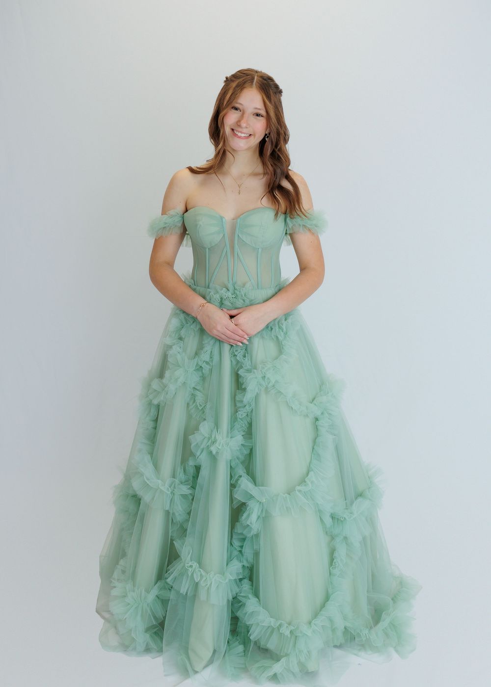 Off-shoulder mint green tulle ball gown with ruffled layers and sweetheart neckline stands against white backdrop.