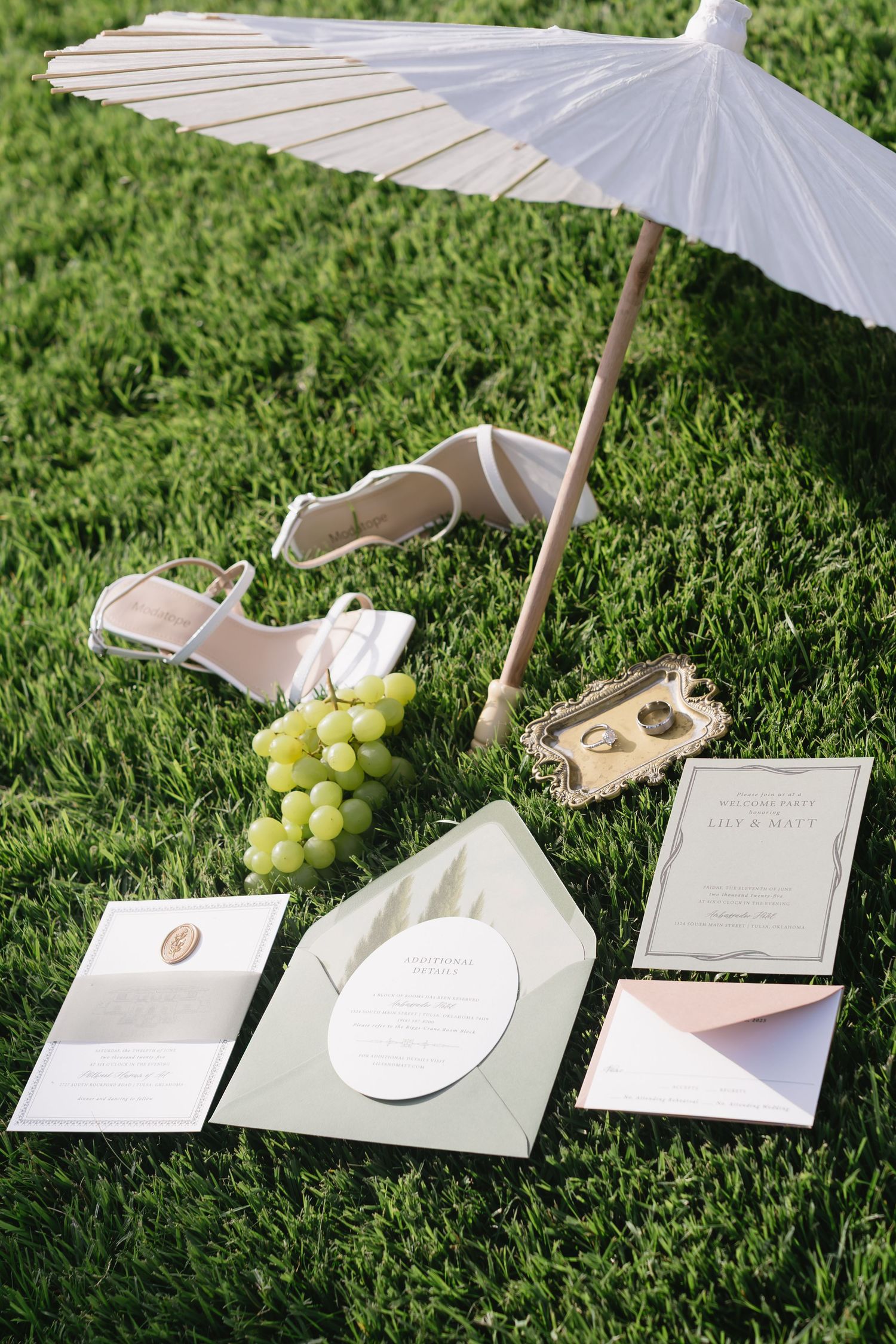 Wedding stationery and accessories arranged on grass with white parasol, sandals, and fresh grapes.