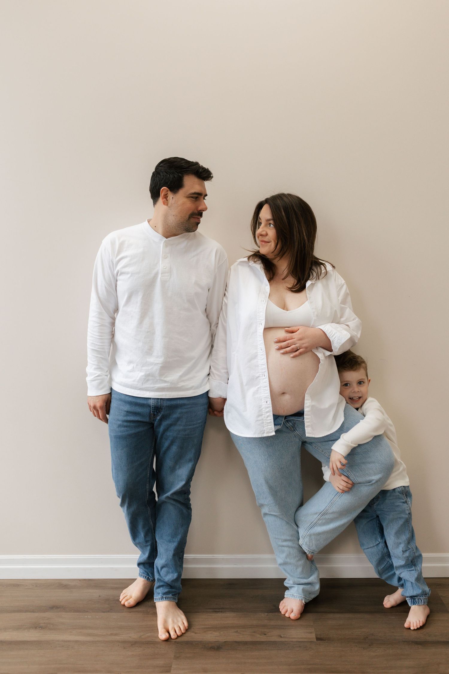 A pregnant woman in a white shirt and jeans poses with her husband and young son against a white wall.